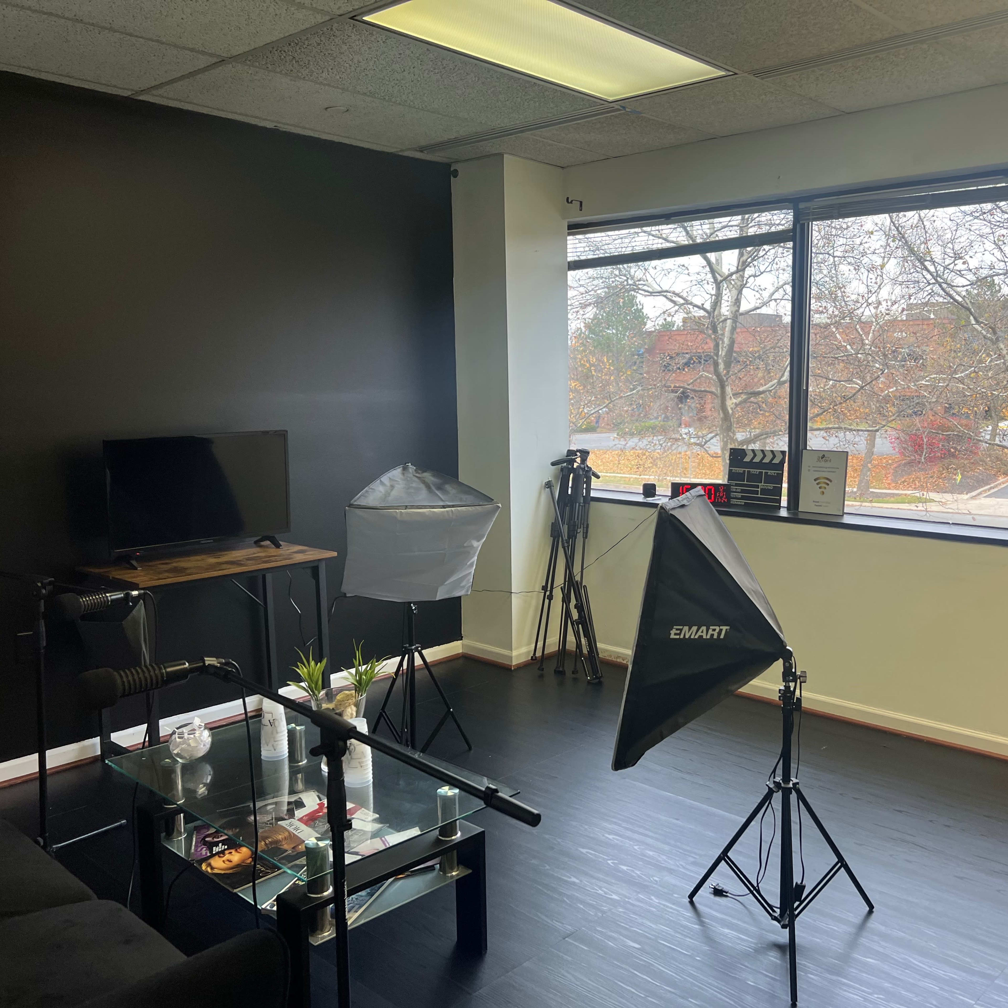 A studio room with black walls, equipped with a television, two light stands, and a window overlooking a tree-lined exterior.