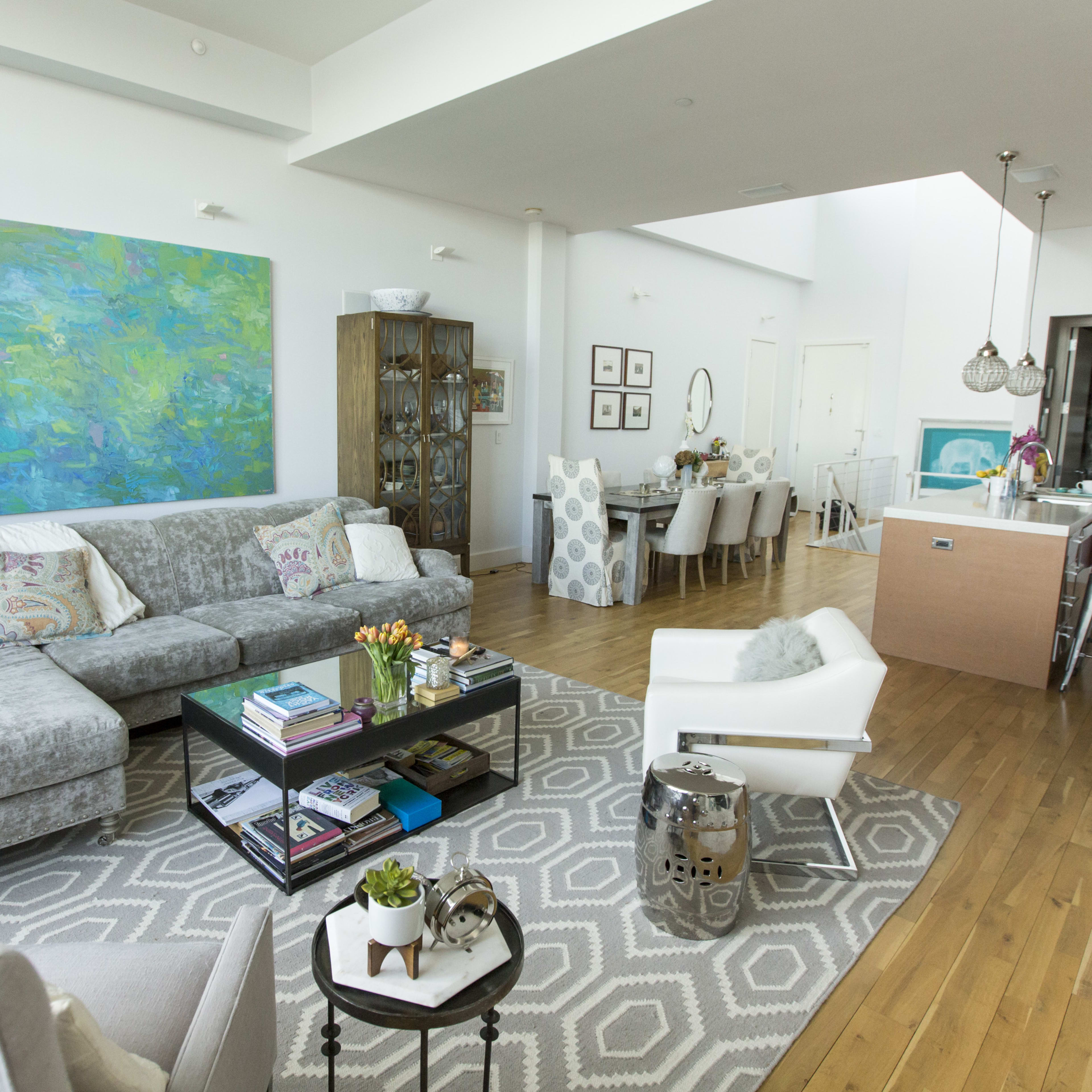A modern living room with a gray sofa, a coffee table filled with books, an art piece on the wall, and a dining area in the background with a table and chairs.