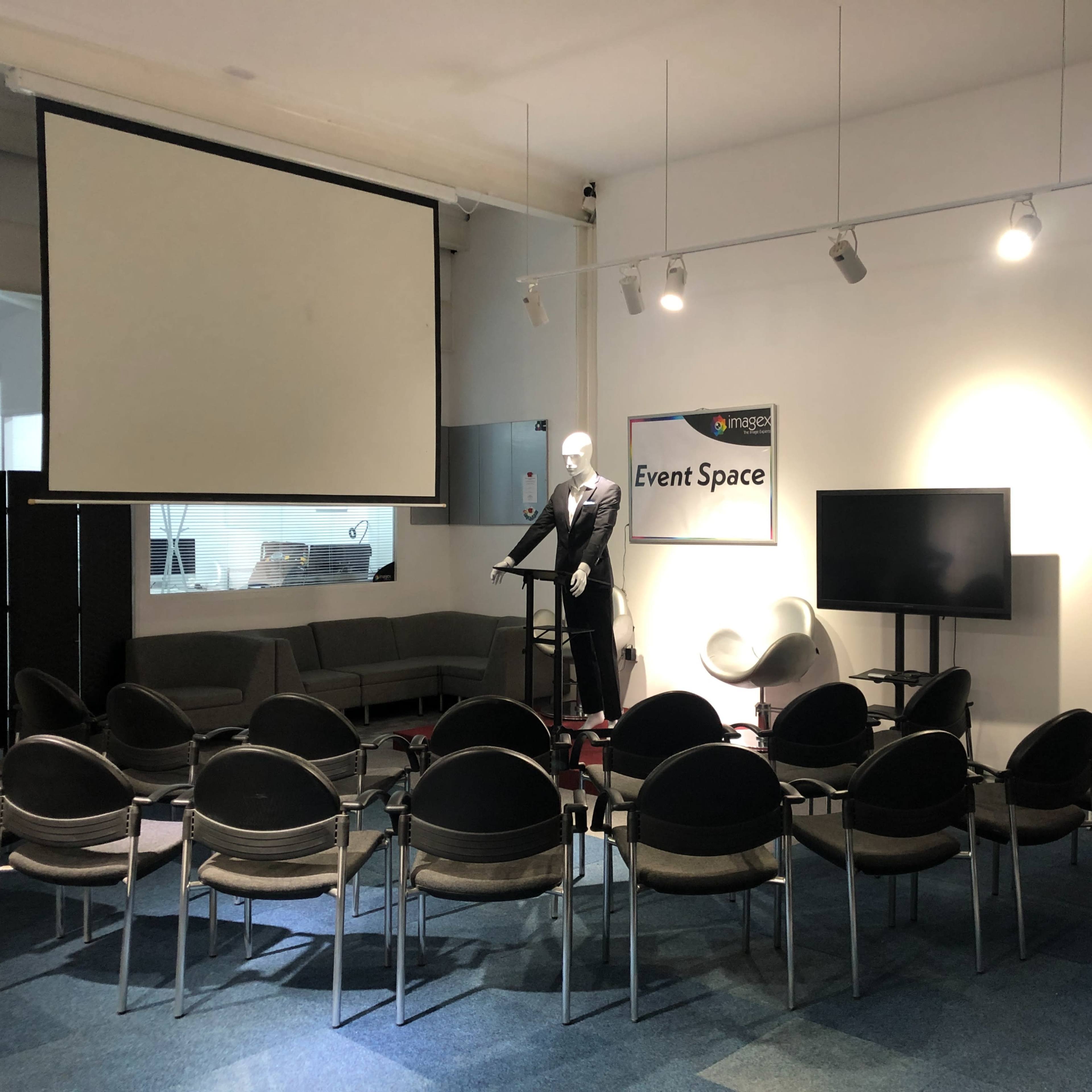 A spacious event space features seating arranged in a semi-circle around a podium, with a projector screen and a television in the background.