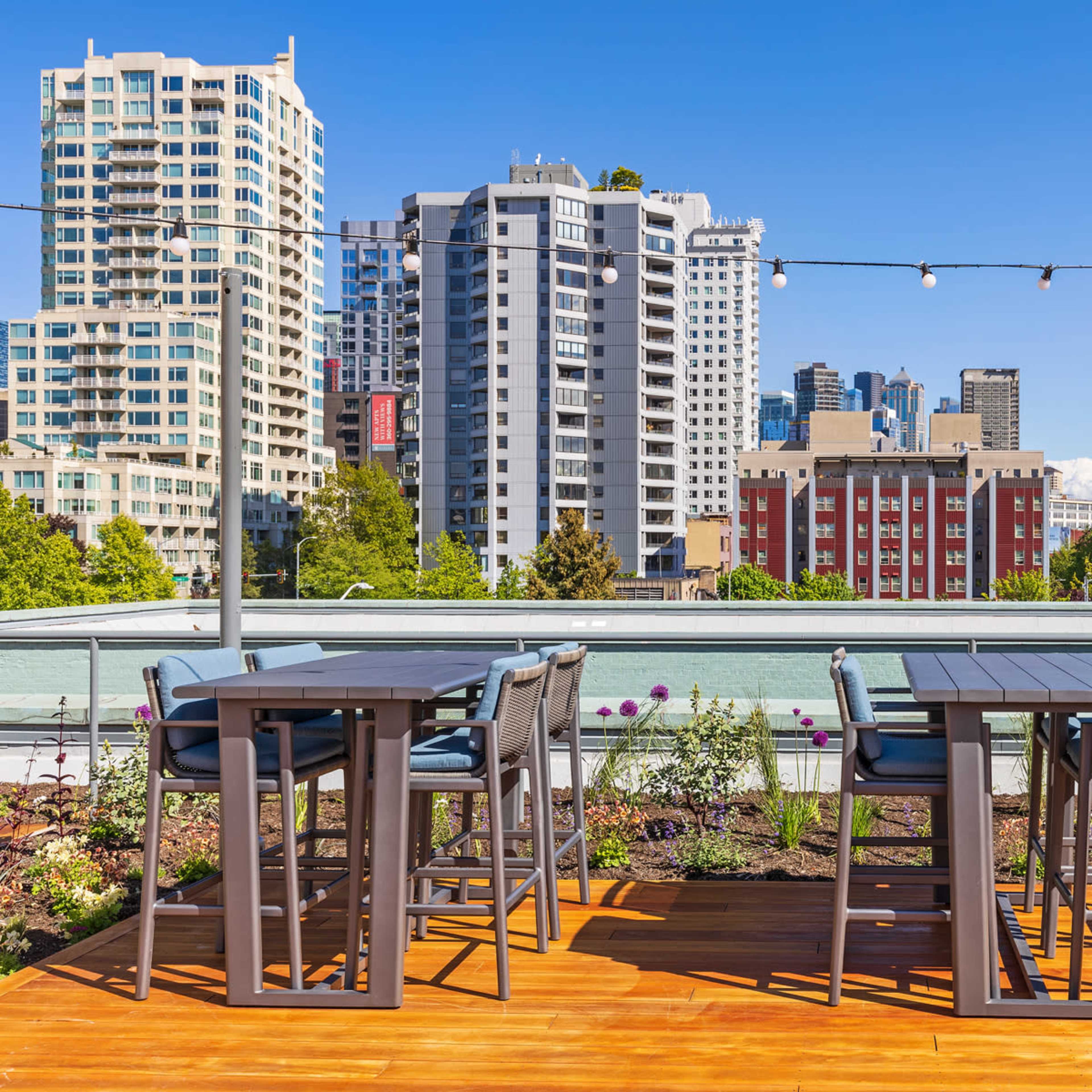 A rooftop terrace with grey furniture and string lights, overlooking a cityscape of tall buildings and clear blue skies.