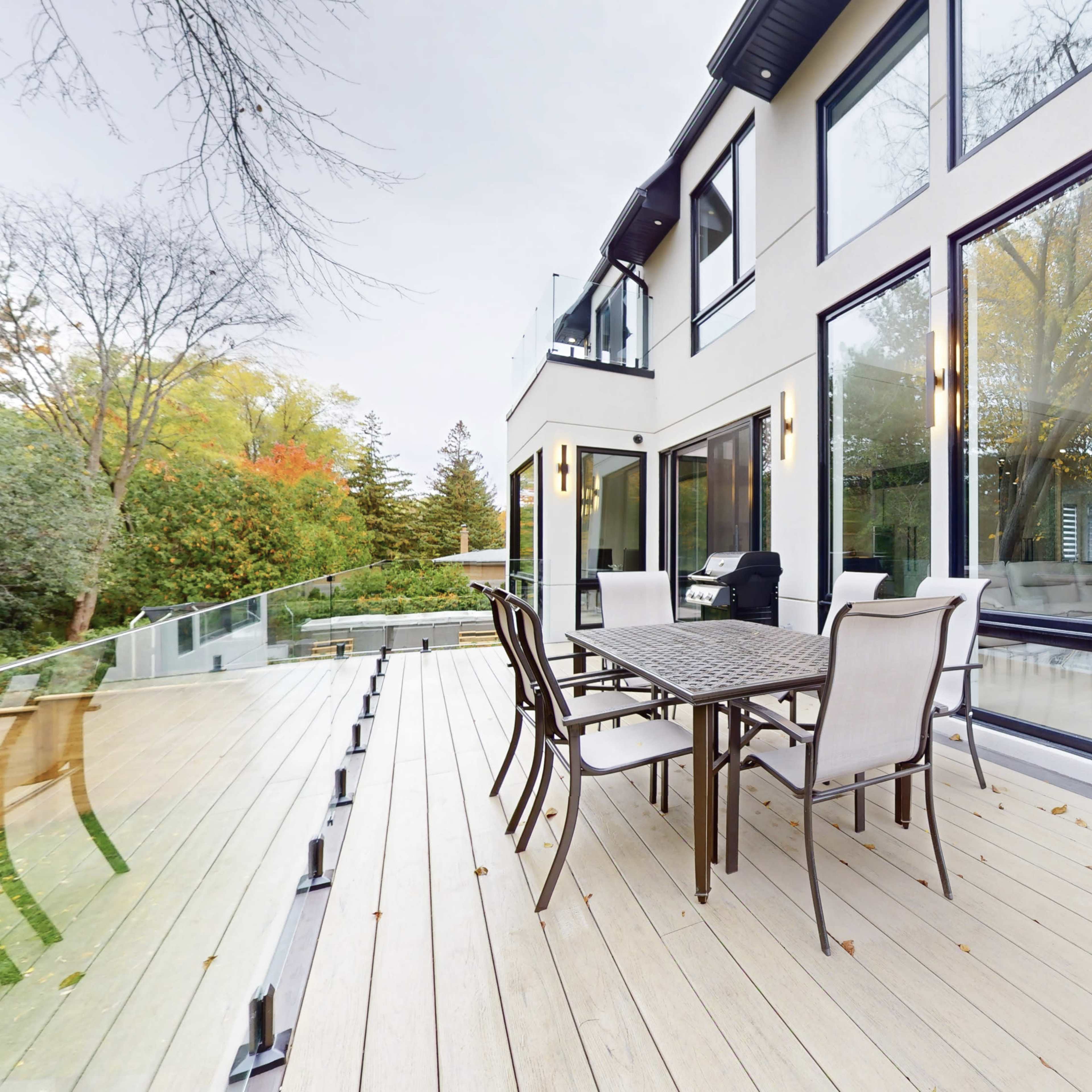 A modern deck with a glass railing features a dining table and chairs, overlooking a landscaped yard and large windows of a contemporary house.