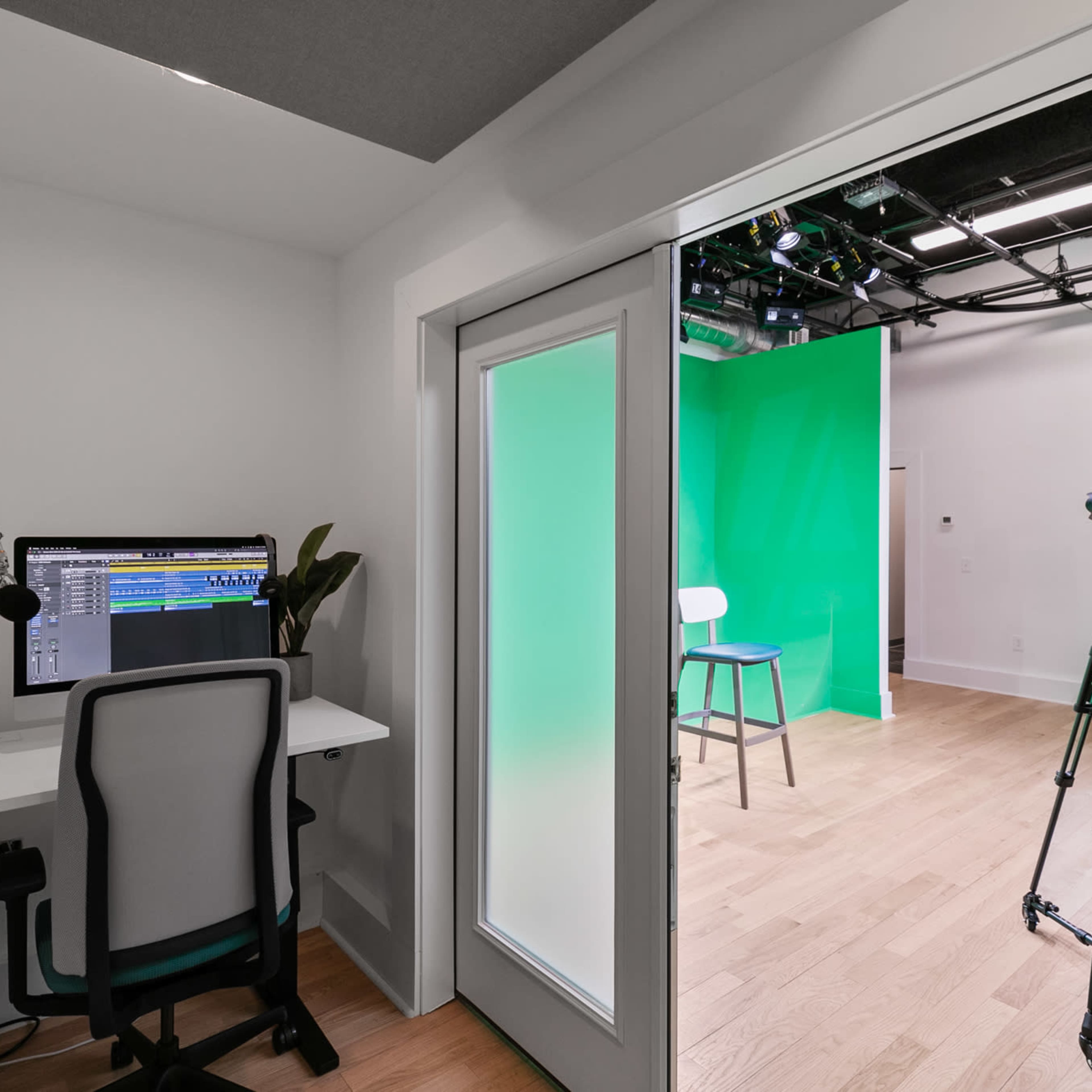 A small room features a desk with a computer and microphone, leading into a studio space with green backdrop and lighting equipment.