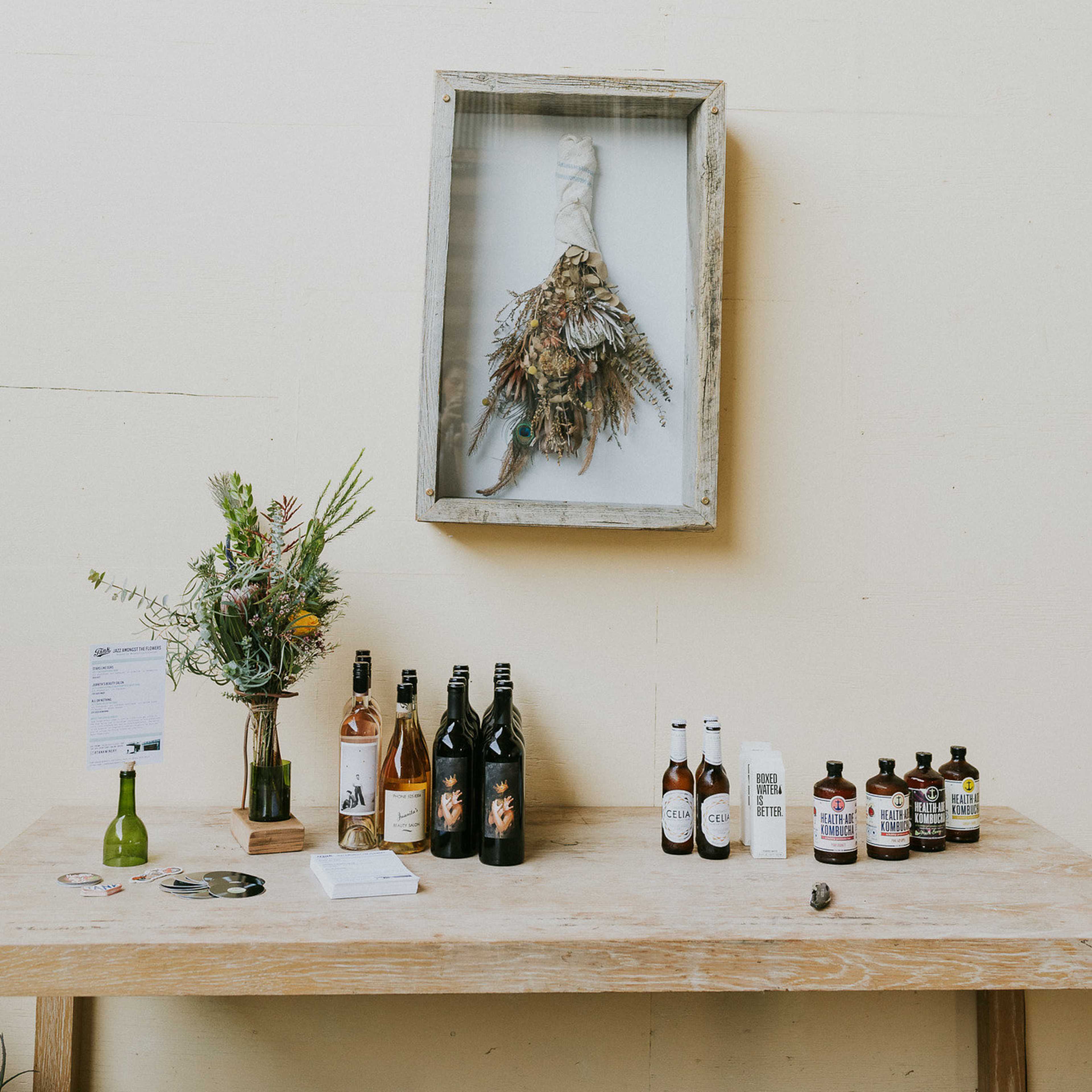A wooden table displays various bottles of beverages alongside a floral arrangement and a framed decoration on the wall.