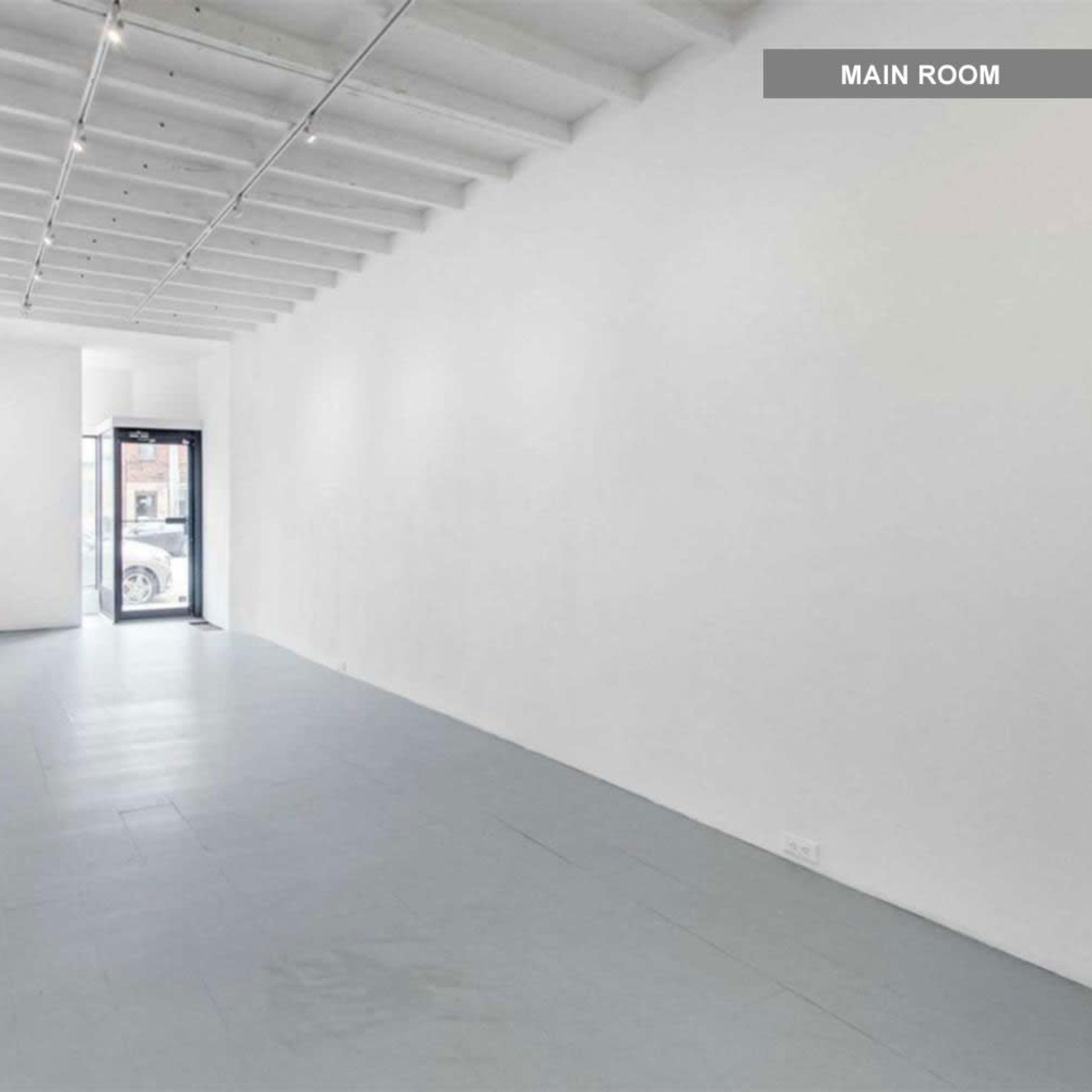 A spacious, empty room with white walls and a light gray floor, leading to a glass entrance.