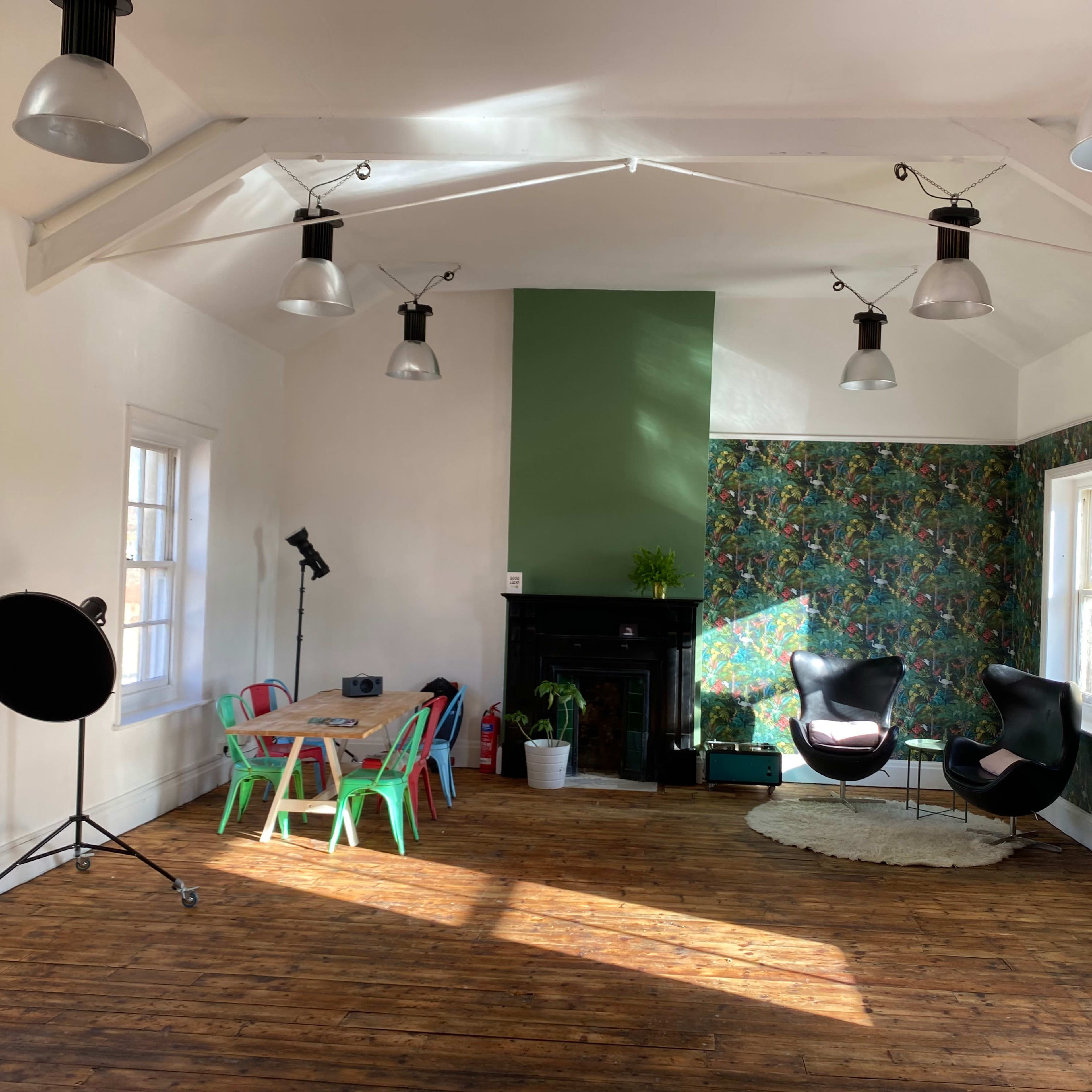 A spacious room with wooden floors, featuring a green accent wall, floral wallpaper, a black fireplace, colorful chairs around a table, and modern light fixtures.