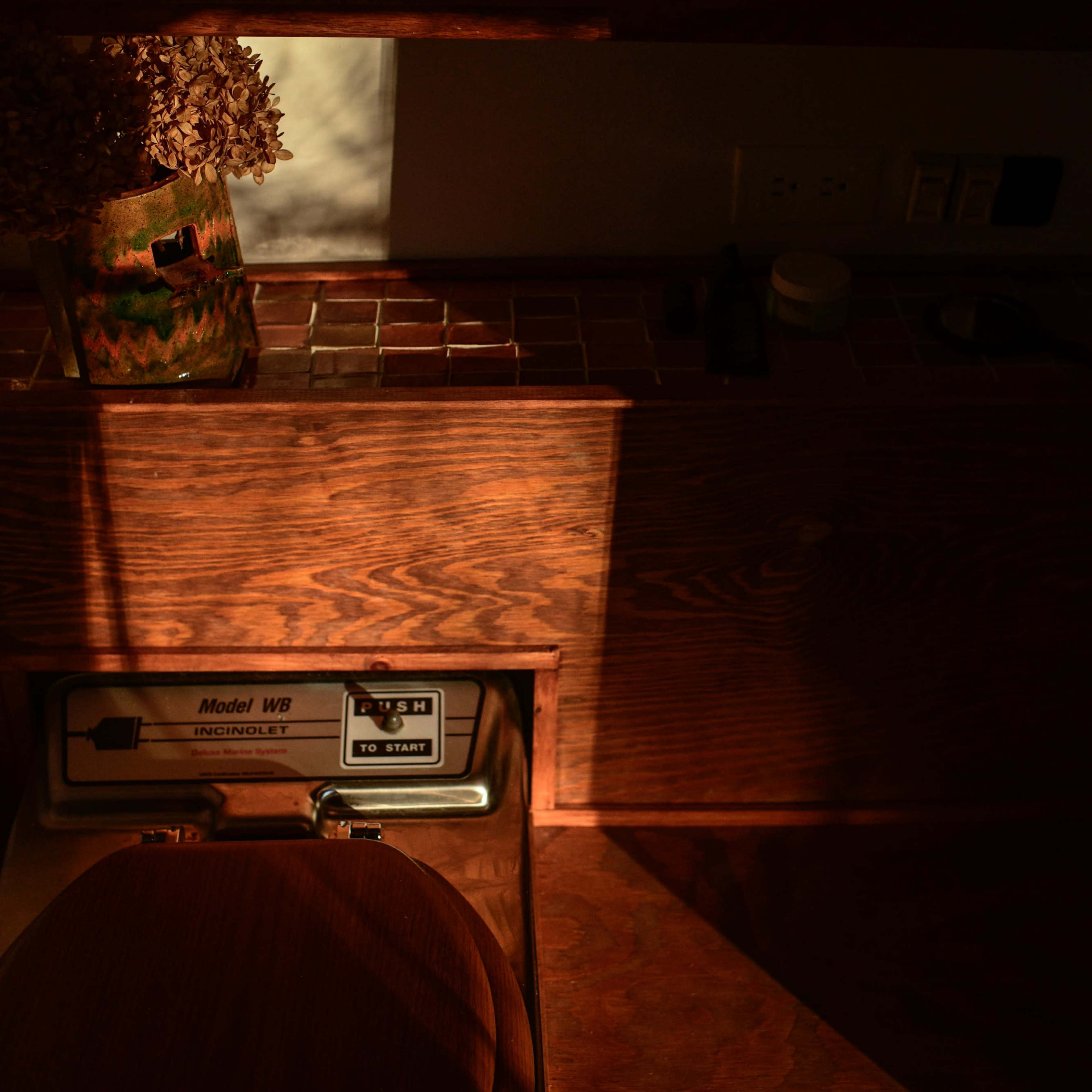 A wooden bathroom setting with a toilet and a flower vase, illuminated by warm sunlight casting shadows across the surface.