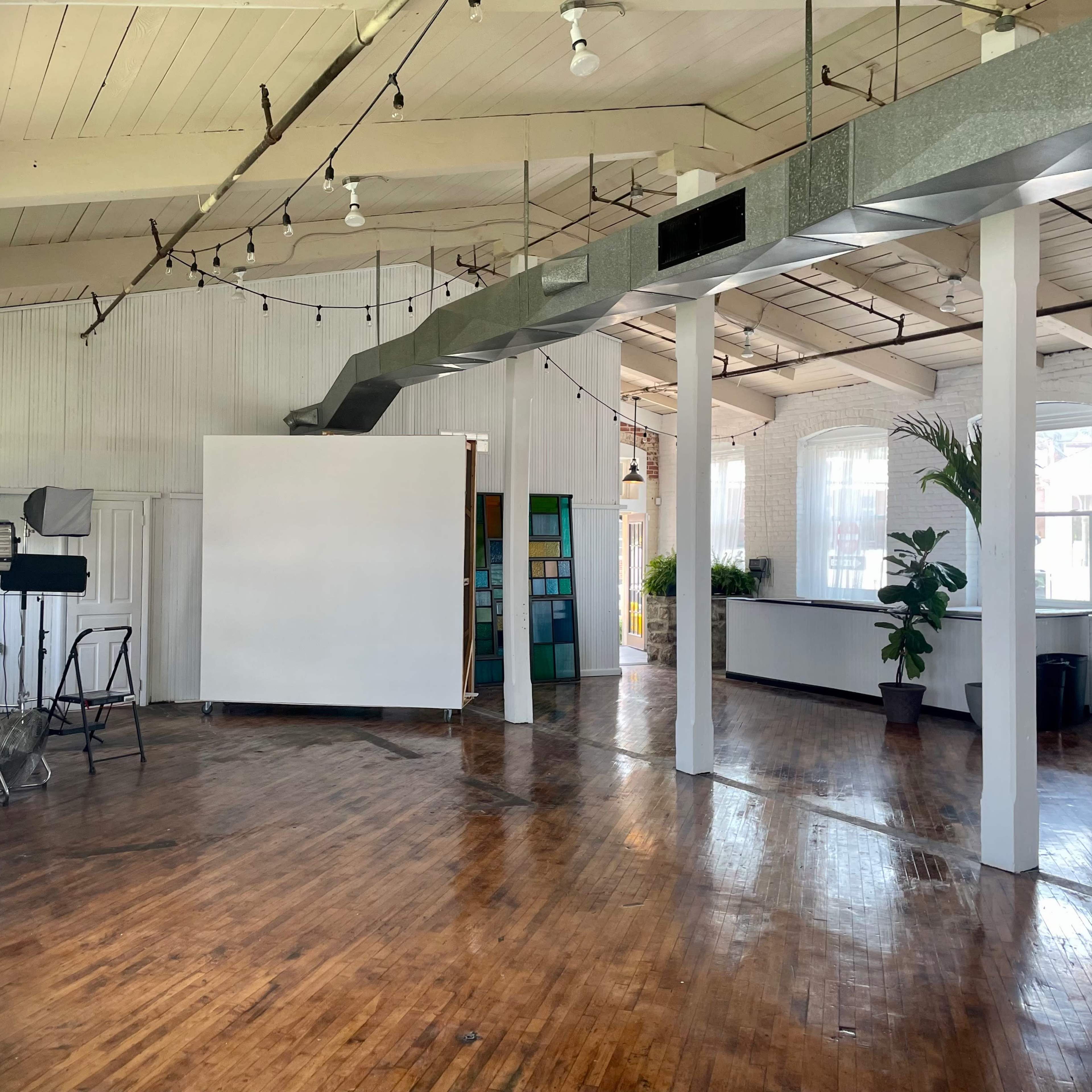 A spacious, well-lit studio with wooden floors, white walls, and large windows, featuring a central blank wall and various studio equipment.