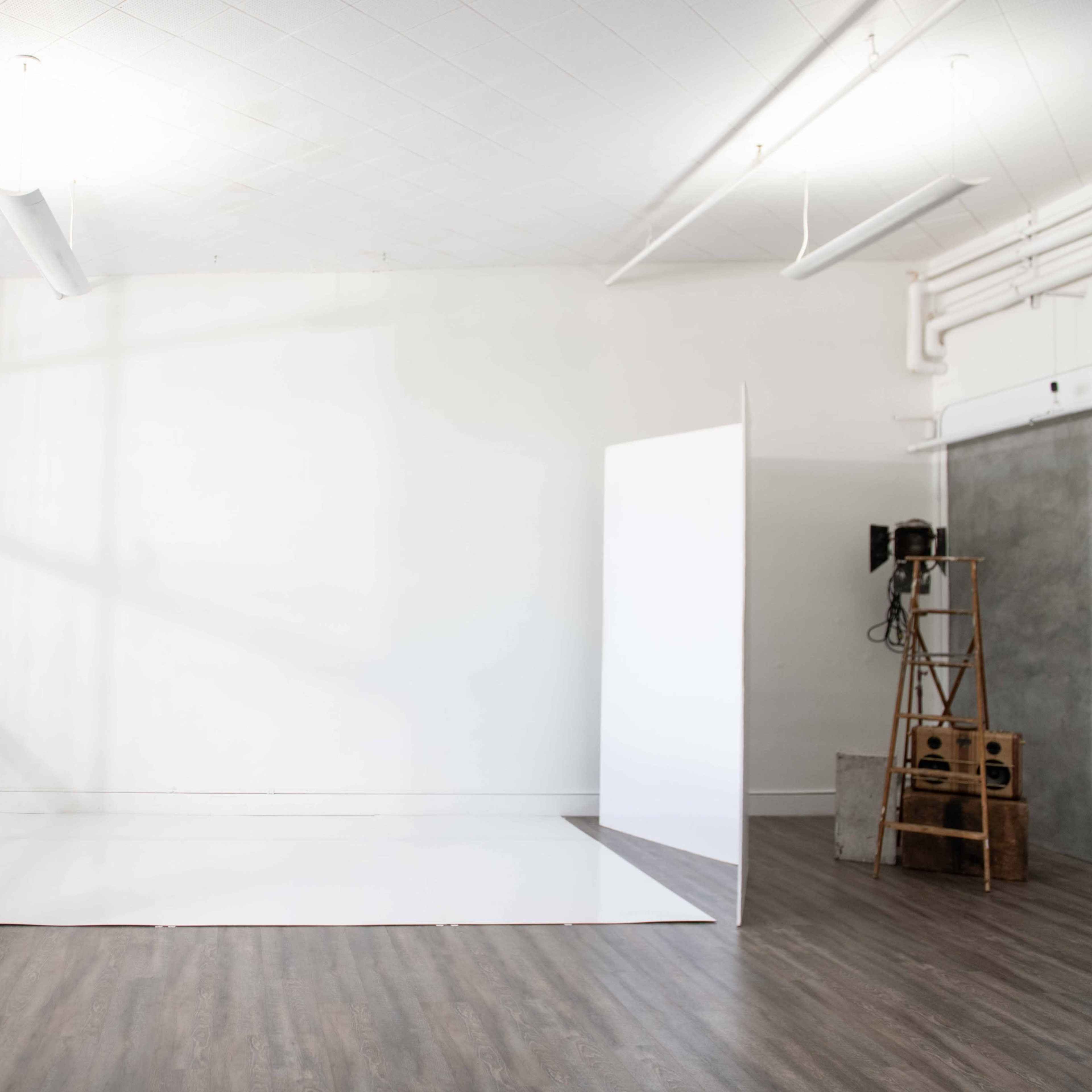 A spacious, empty room with a white wall and a large white floor panel, illuminated by natural light coming through large windows.
