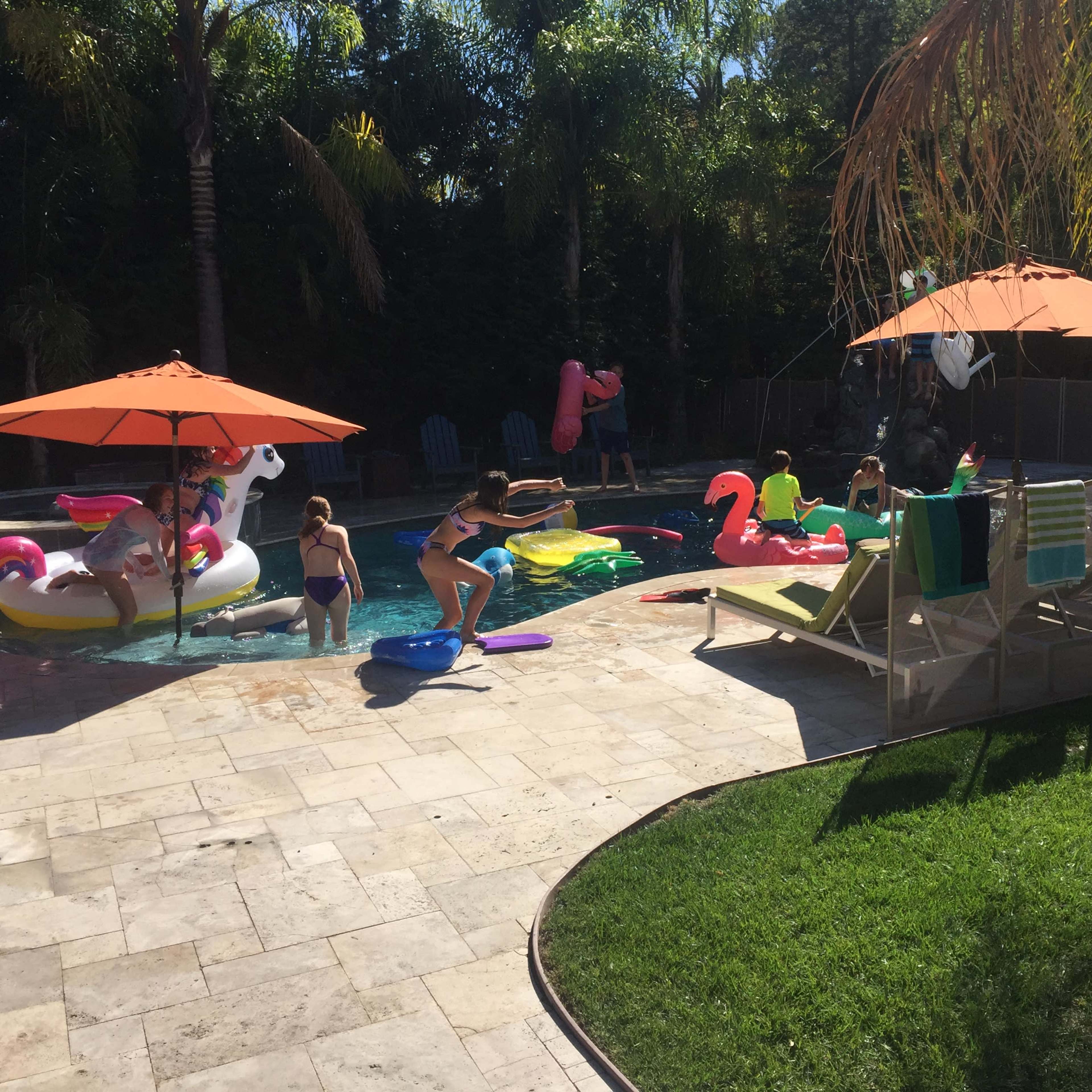 A group of children enjoys playing in a backyard pool surrounded by colorful floaties, lounge chairs, and sun umbrellas.