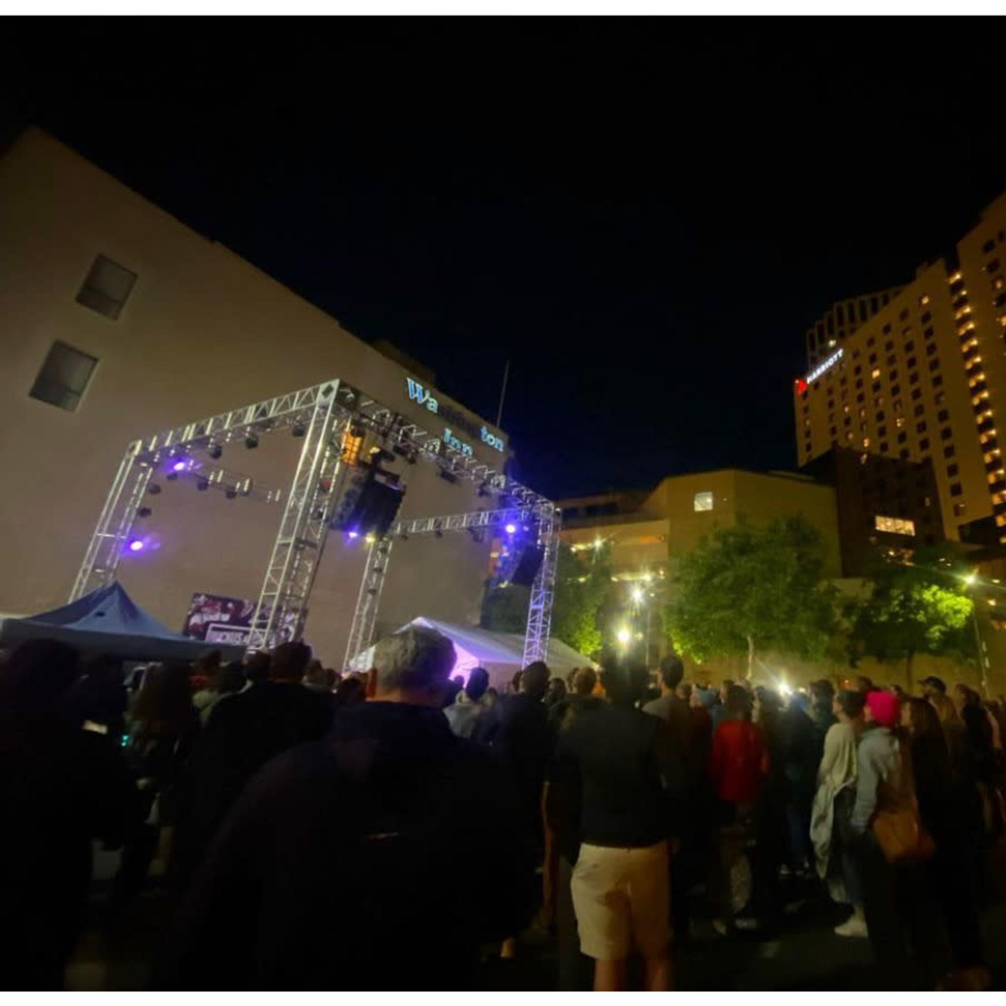 A crowd gathers around a stage illuminated by lights at an outdoor event in a city at night.