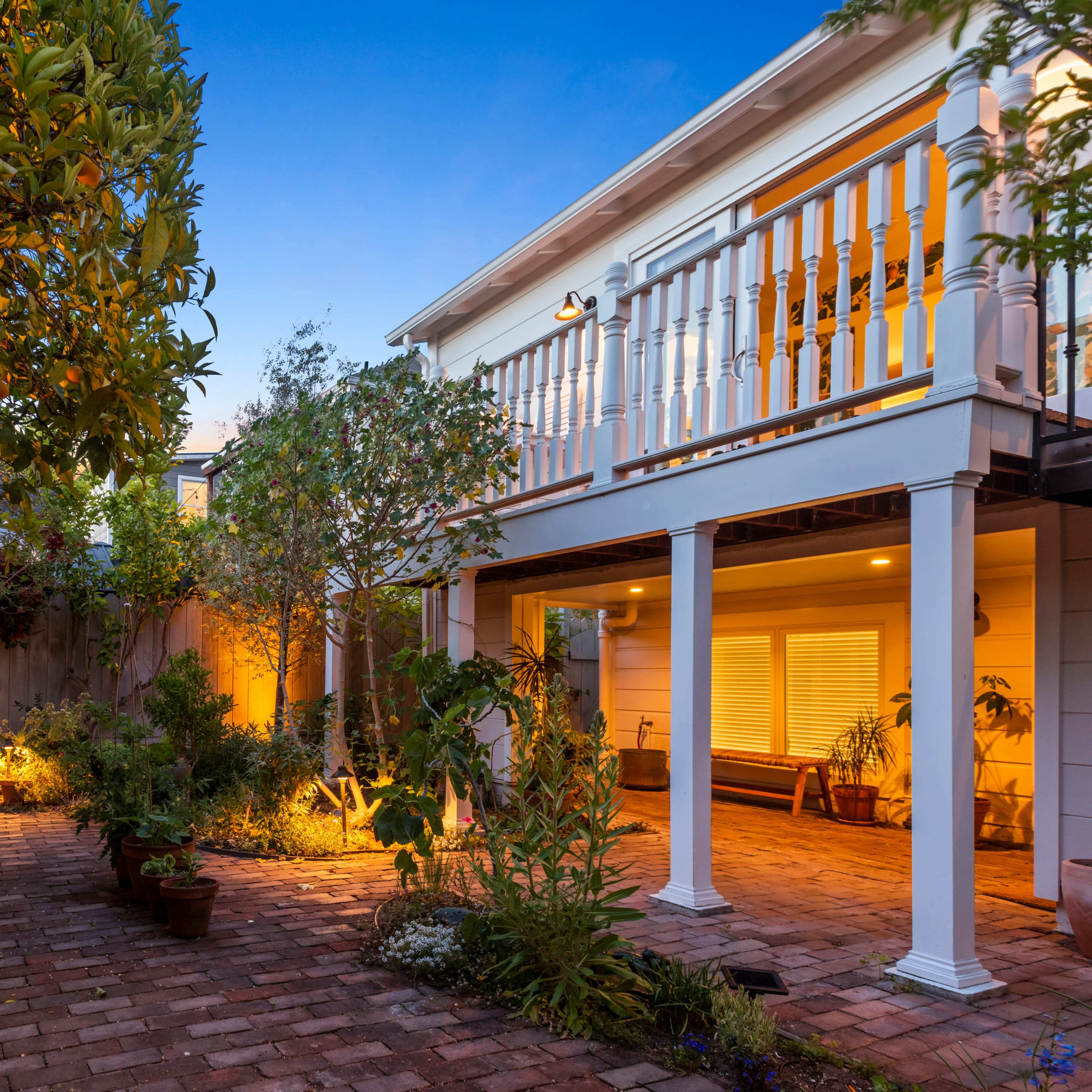 A brick-paved backyard with garden plants, two white chairs, and a lit area beneath a deck.