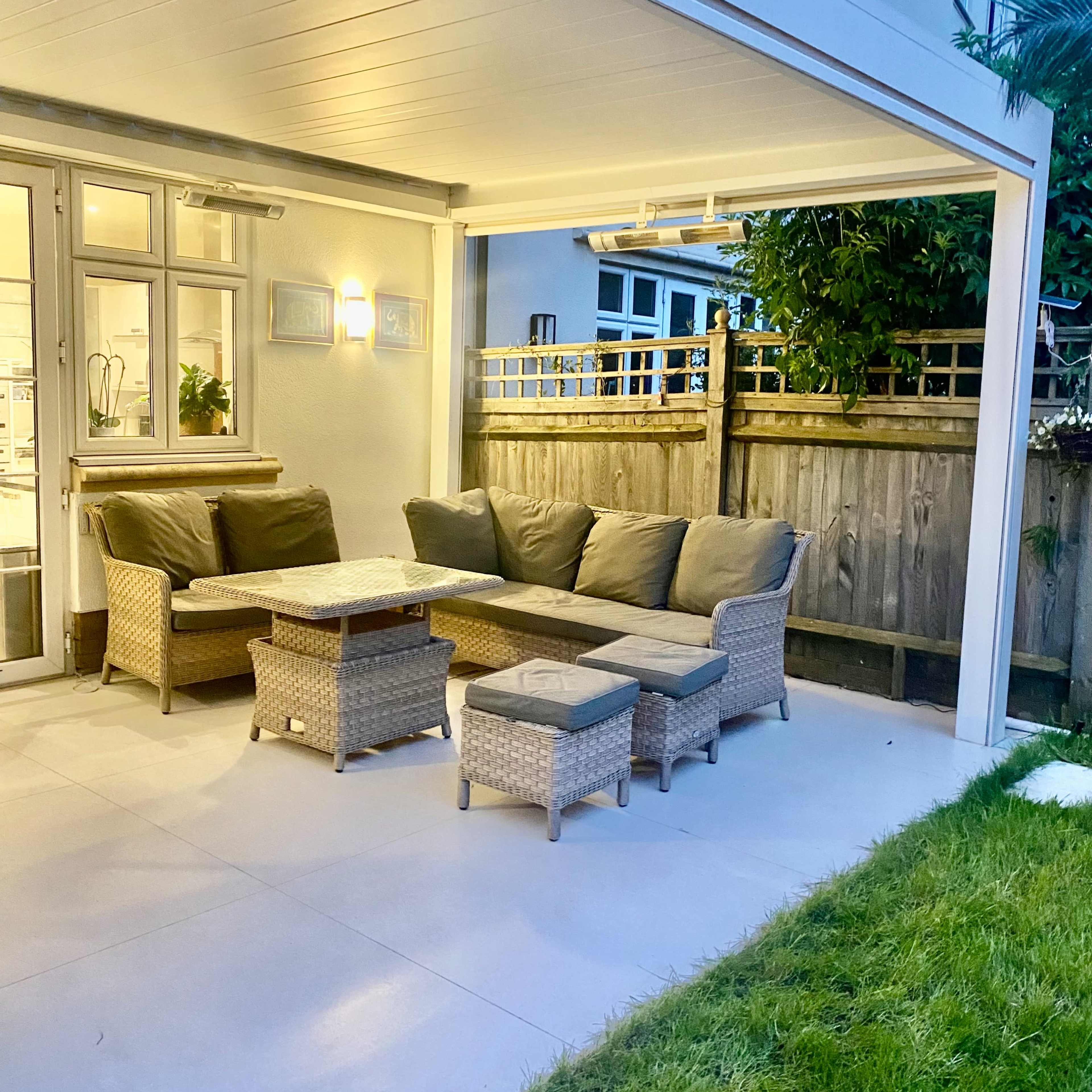 A cozy outdoor seating area featuring a wicker sofa set with cushions and a rectangular table, situated under a covered patio next to a lawn.