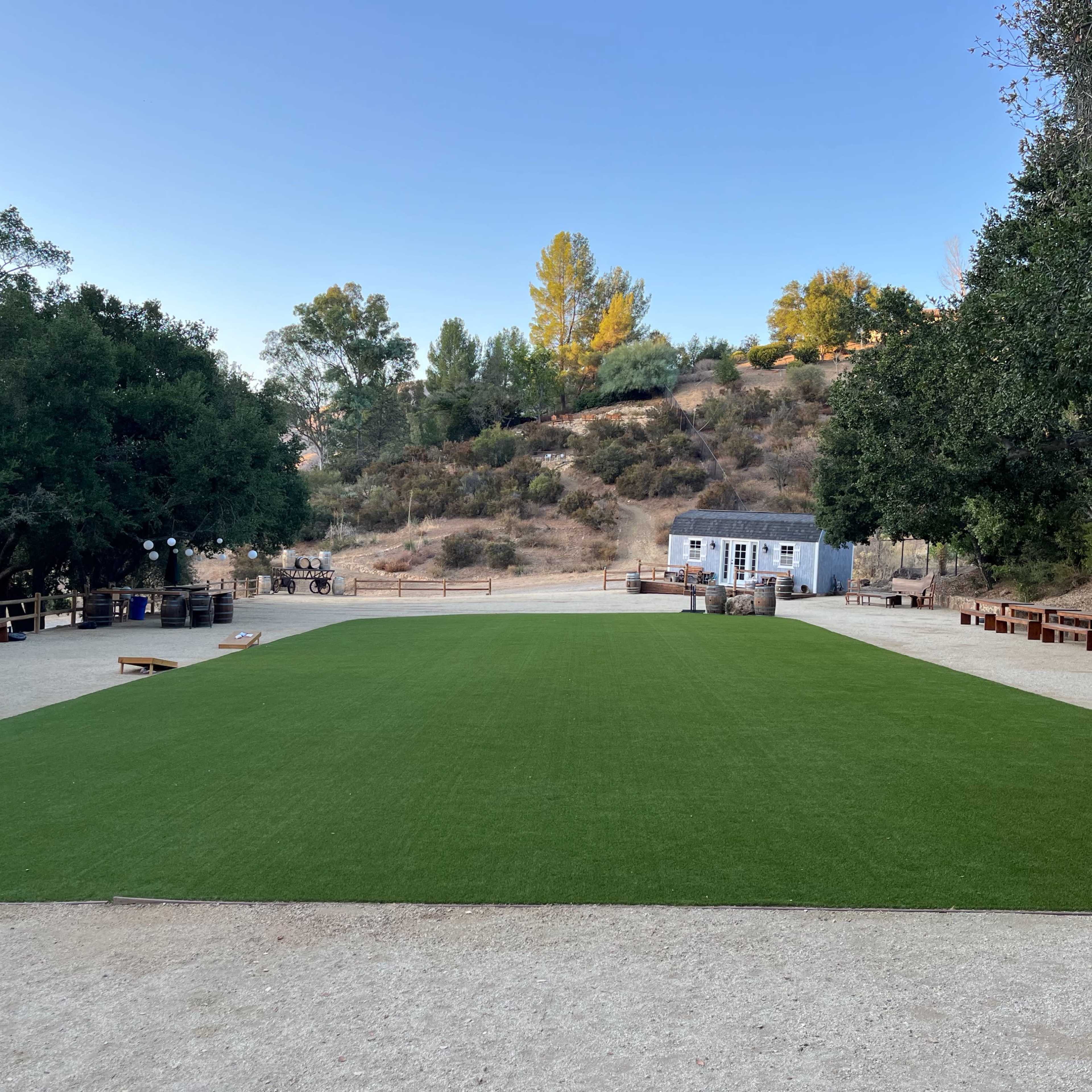 A rectangular area of artificial grass is surrounded by trees and rustic wooden furniture in a rural setting.