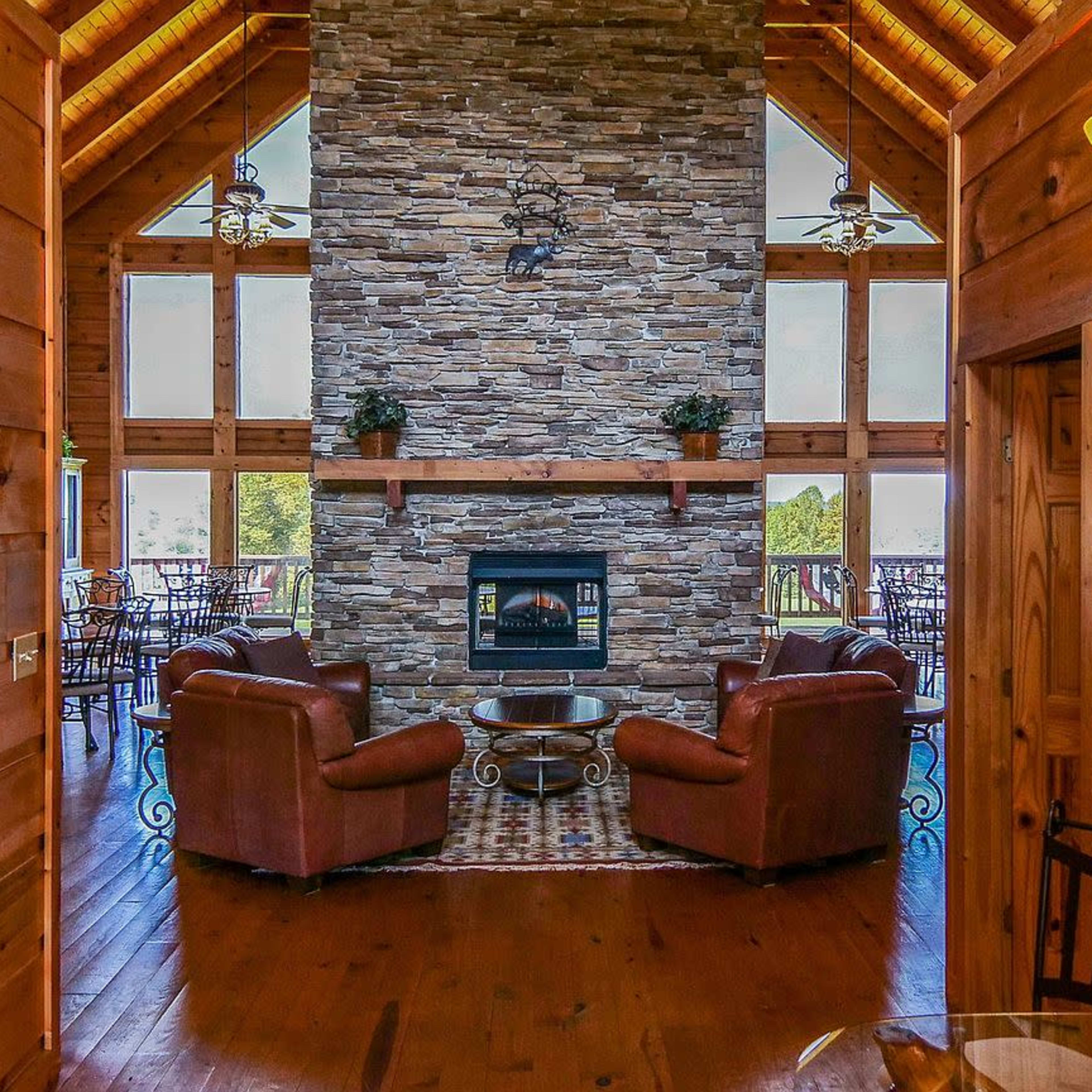A wooden interior of a cabin with a stone fireplace at its center, flanked by large windows and wooden furniture.