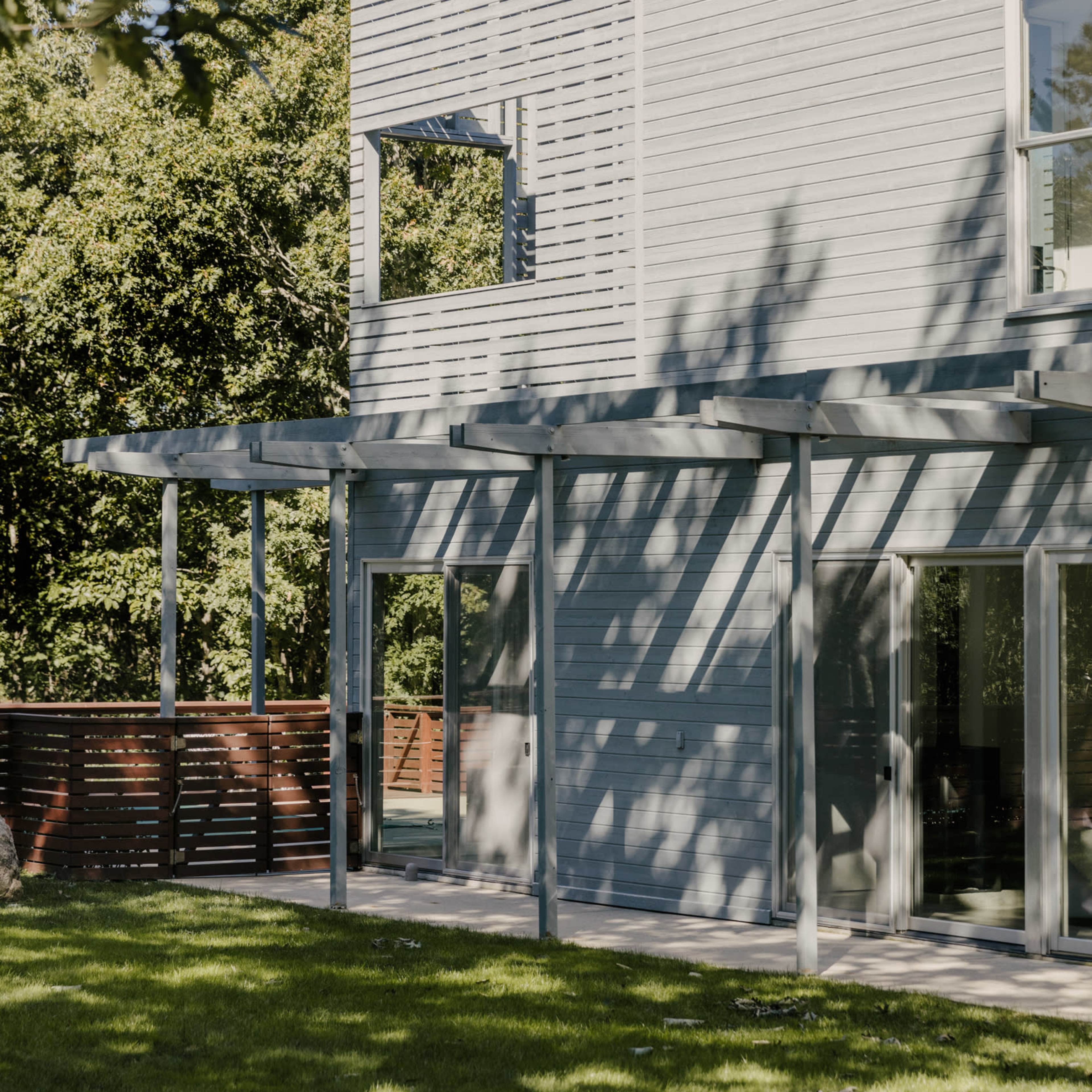 A modern light-colored house with large windows and a covered patio, situated in a grassy yard surrounded by trees.