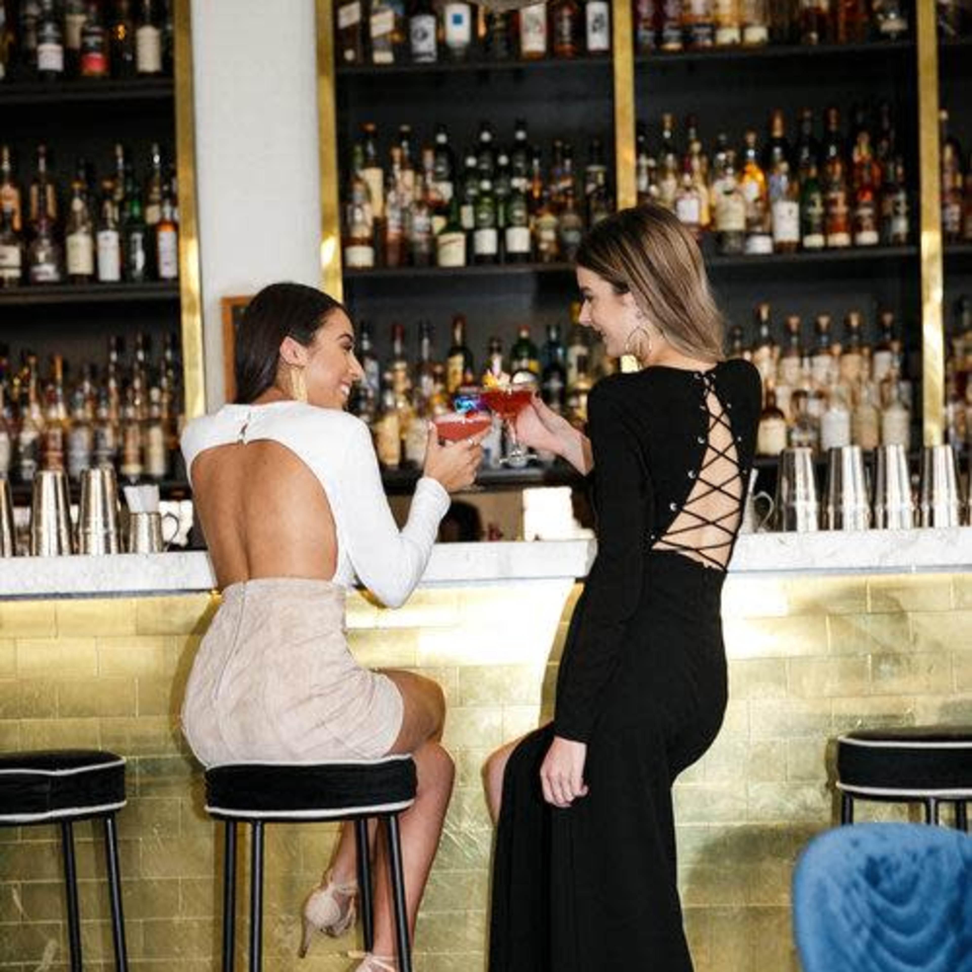 Two women in stylish evening dresses clink cocktail glasses while seated at a bar with a well-stocked shelf of liquor behind them.