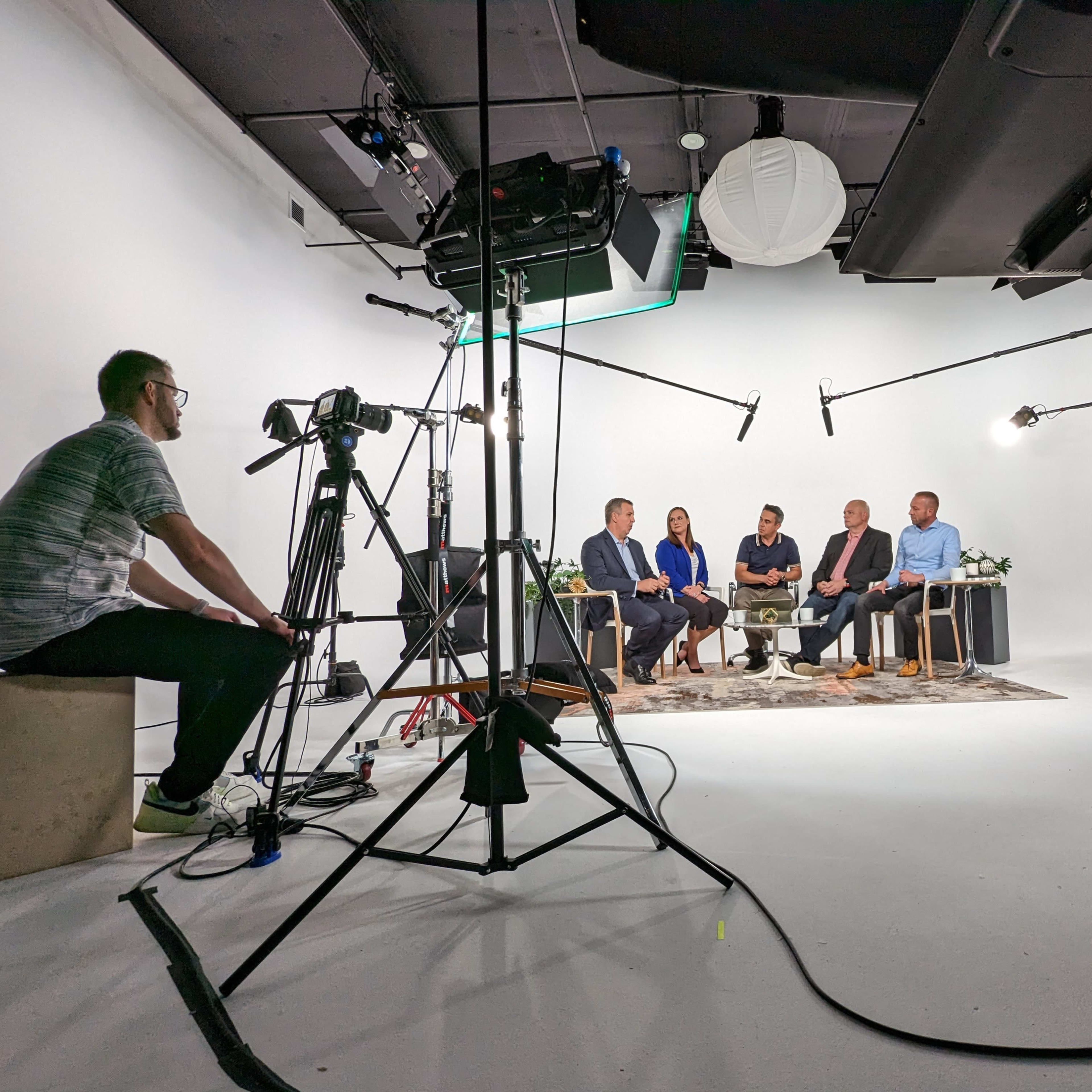 A film crew sets up a discussion panel with five participants on a white backdrop in a studio, surrounded by various cameras and lighting equipment.