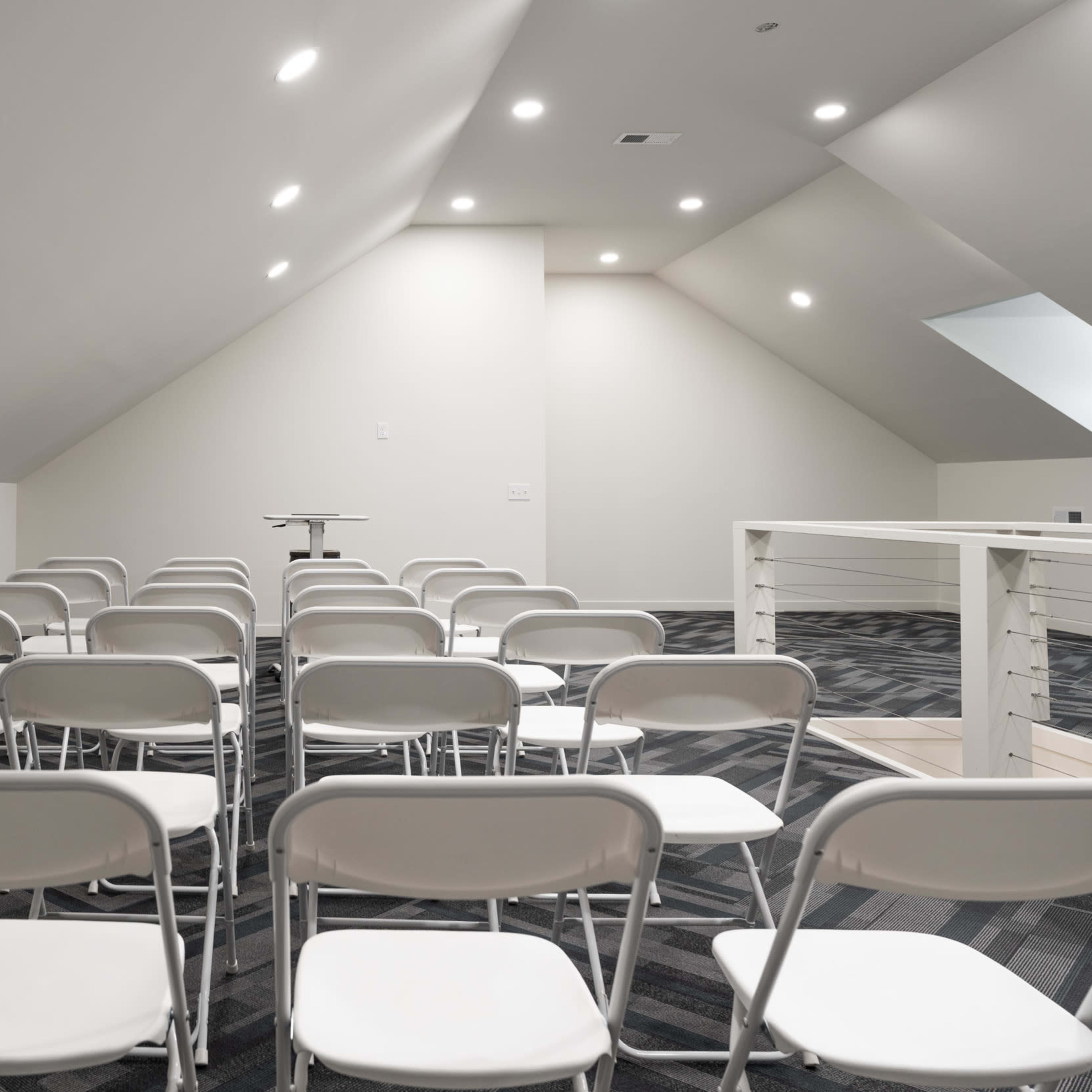 A room with sloped ceilings arranged with several rows of white folding chairs facing a central table.