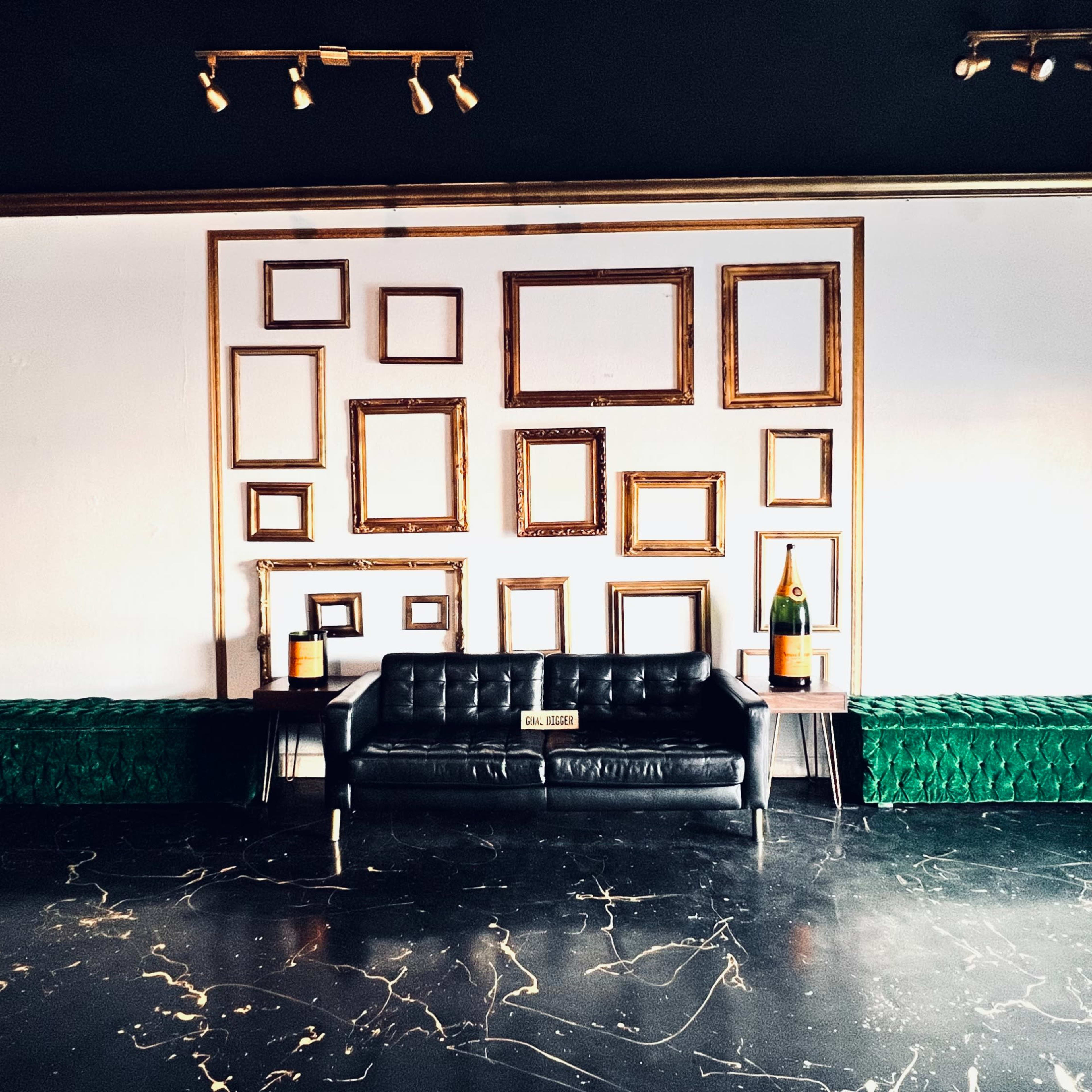 The scene features a black leather sofa positioned between two green storage benches, against a wall decorated with an arrangement of empty wooden frames.