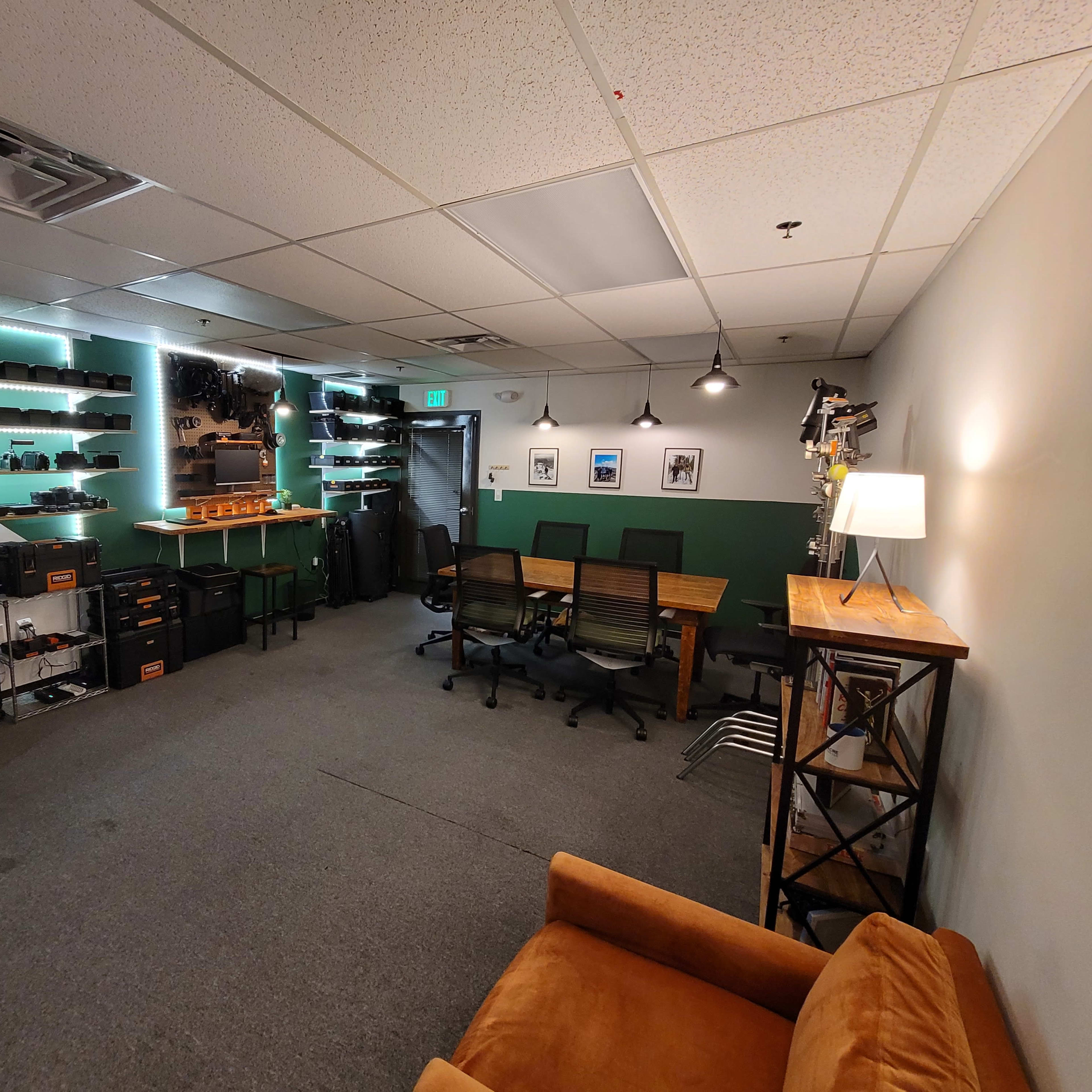 A modern office space with green accent walls, equipped with shelves holding equipment, several chairs around a table, and a cozy seating area featuring an orange couch.