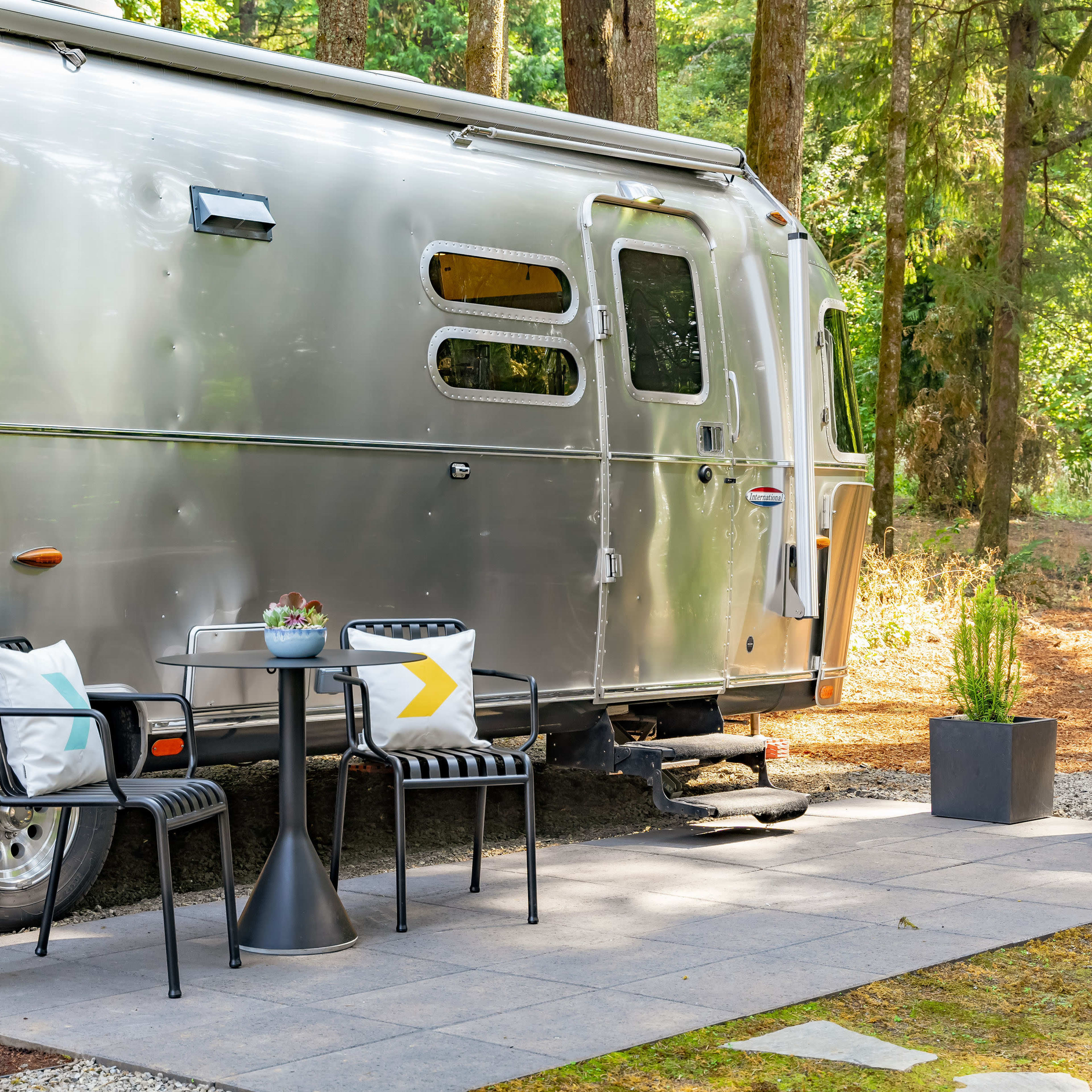 An aluminum Airstream trailer is parked beside a stone patio featuring a small table and chairs, surrounded by trees.