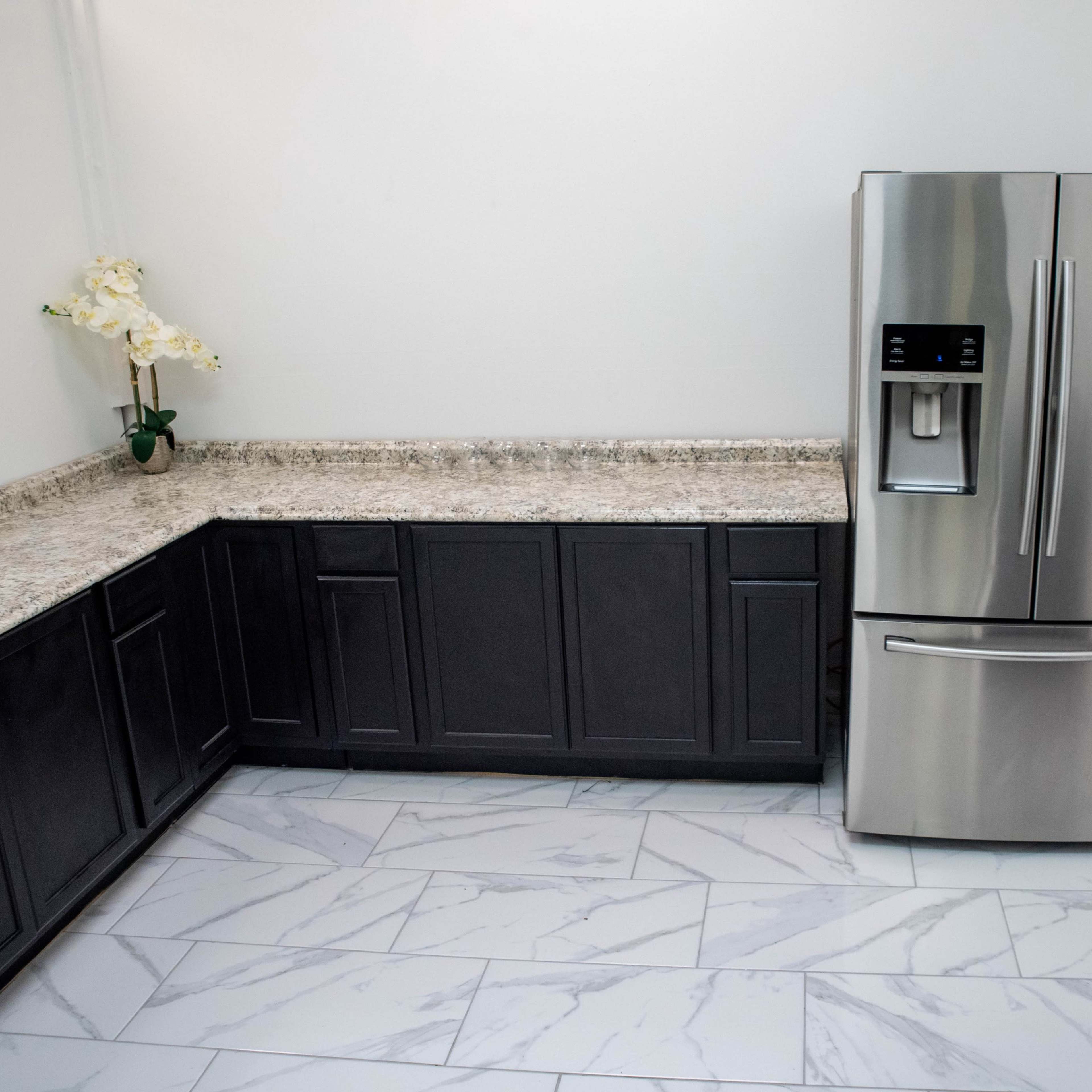 A modern kitchen with dark cabinetry, a stainless steel refrigerator, and a black trash bin on a tiled floor.