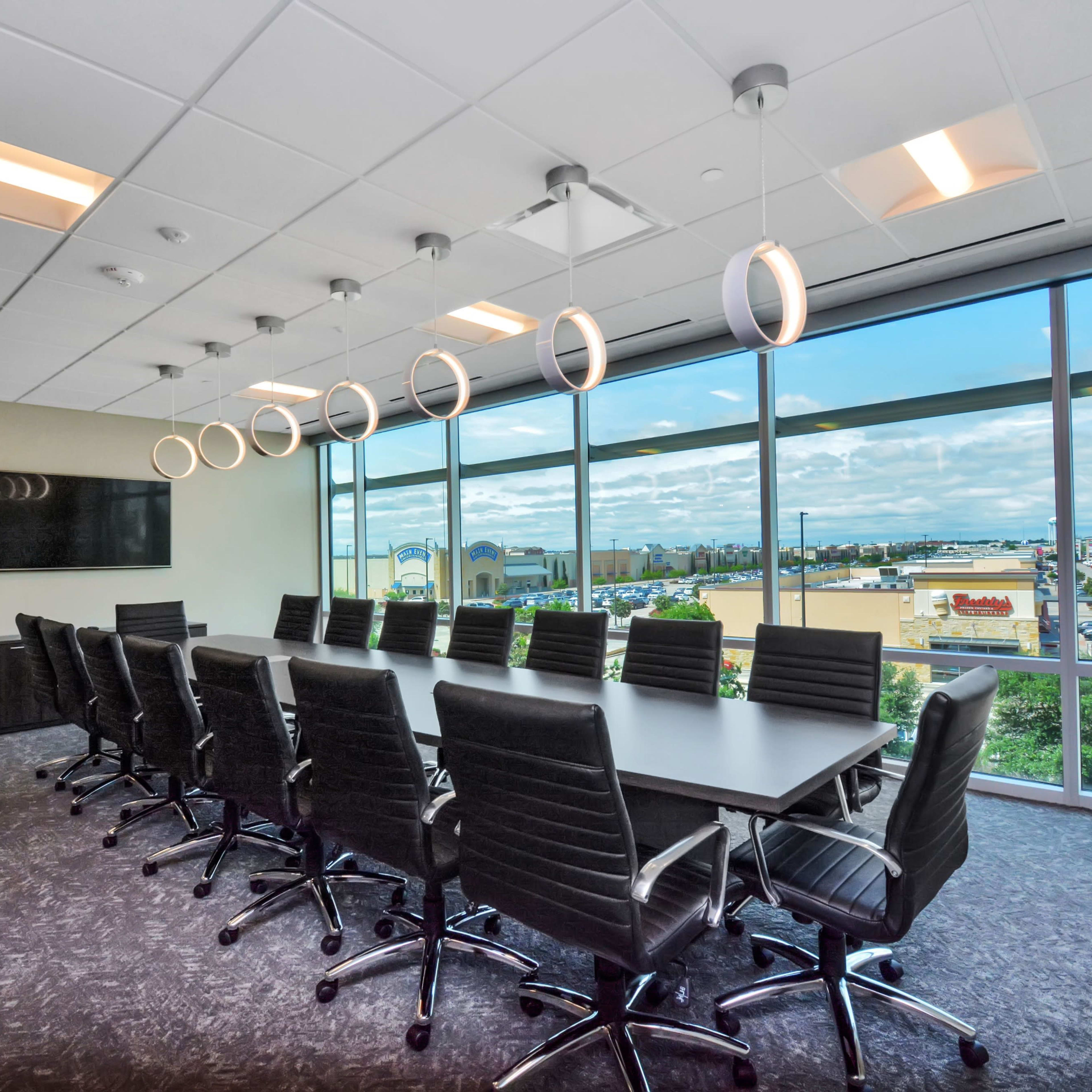 A large, modern conference room features a long black table surrounded by ergonomic chairs, with floor-to-ceiling windows providing a view of the outside.