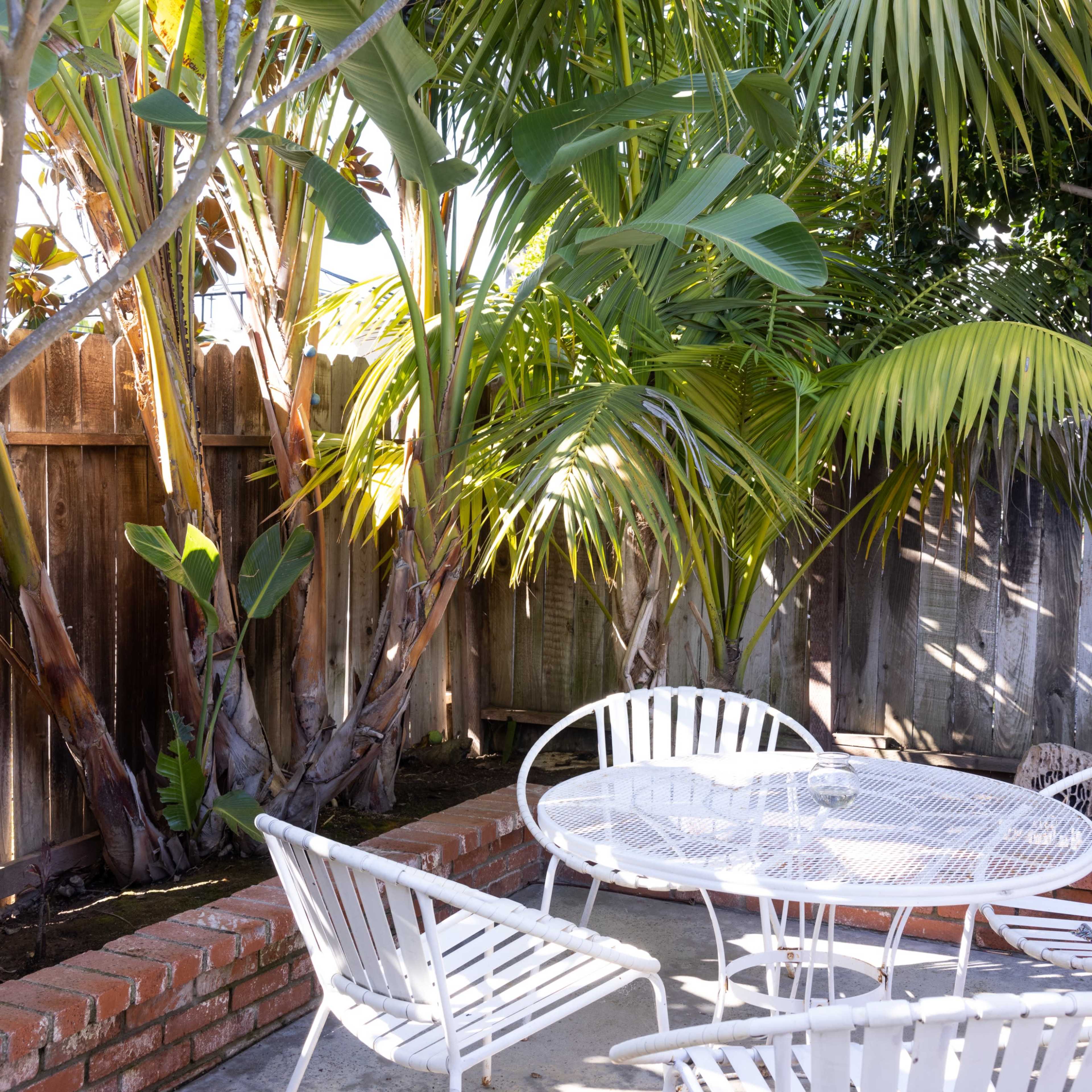 A patio area features a round table surrounded by white chairs, nestled among tall palm trees and a wooden fence.