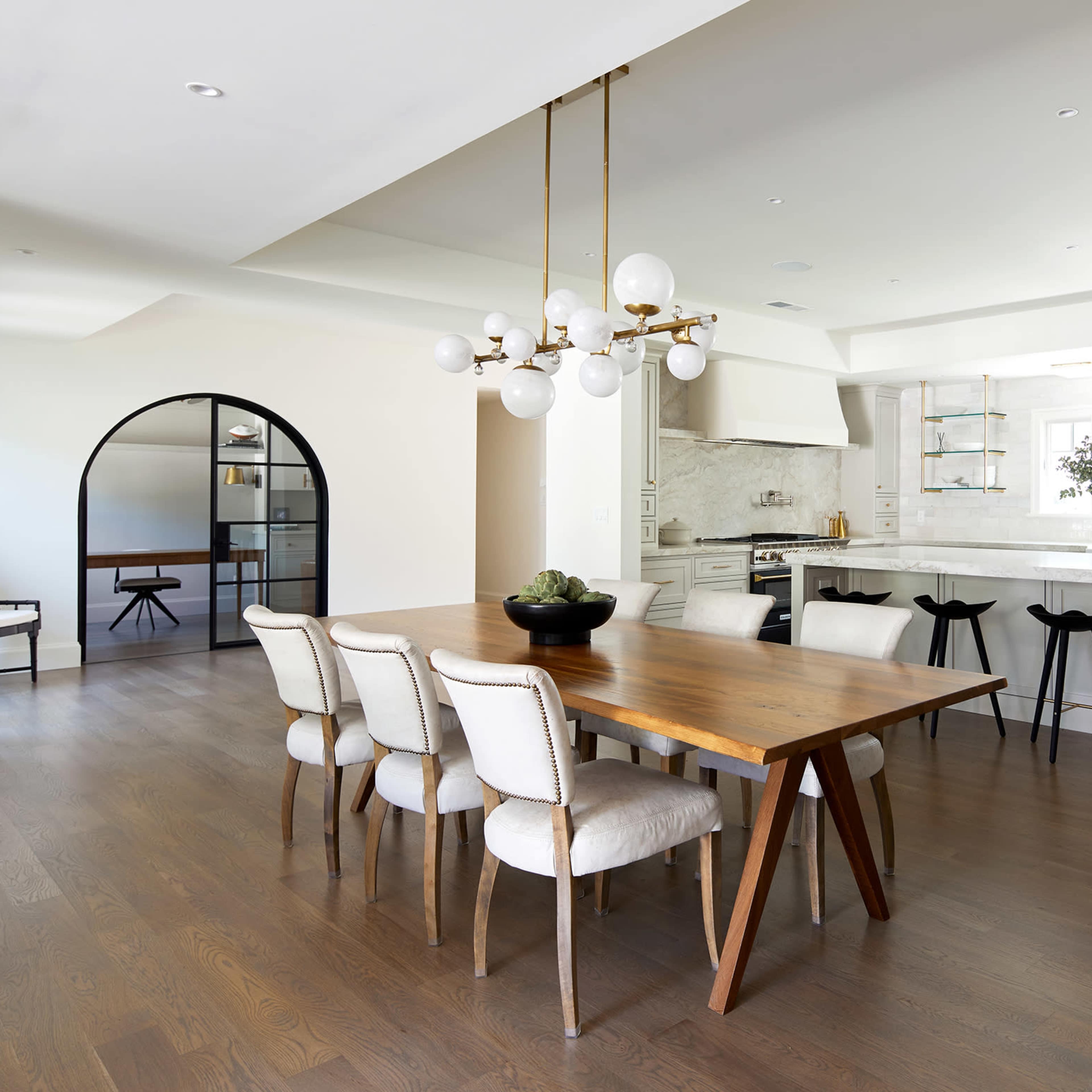 A modern dining area with a wooden table surrounded by six upholstered chairs and a large kitchen in the background, complete with white cabinetry and a sleek island.