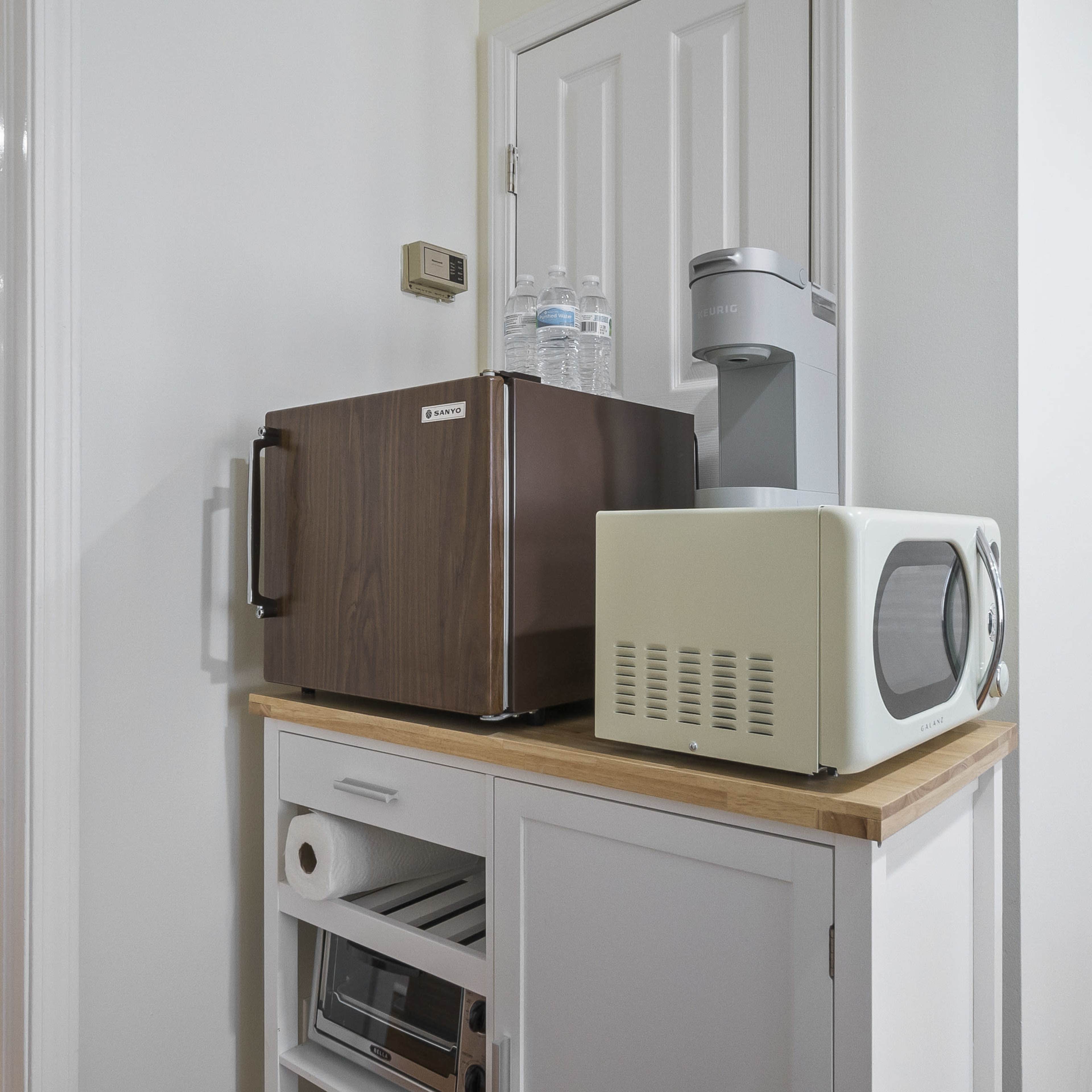 A compact kitchenette area features a small refrigerator, a microwave, and a water dispenser on a wooden countertop.