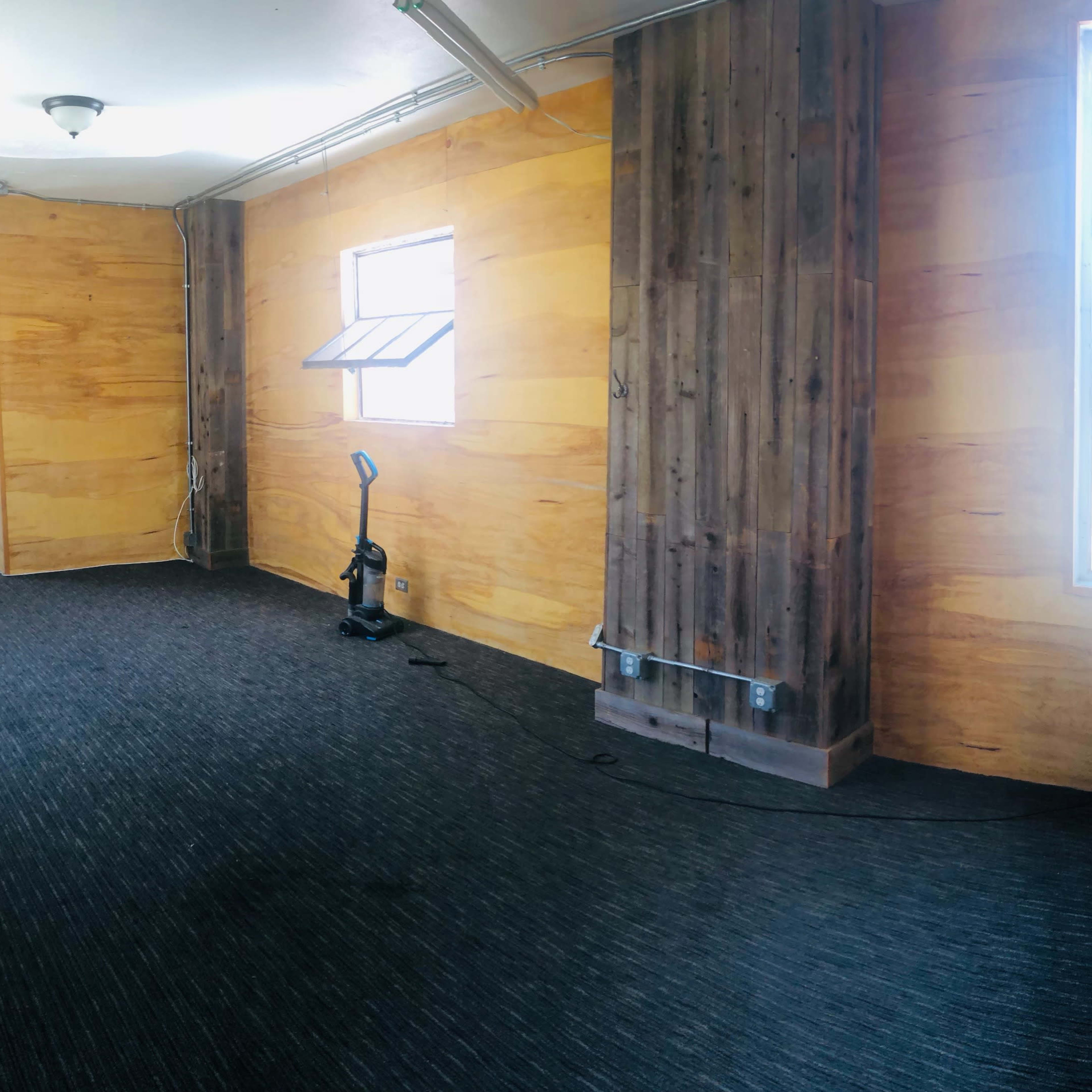 An empty room with wooden walls, a window, and a vacuum cleaner in the corner.