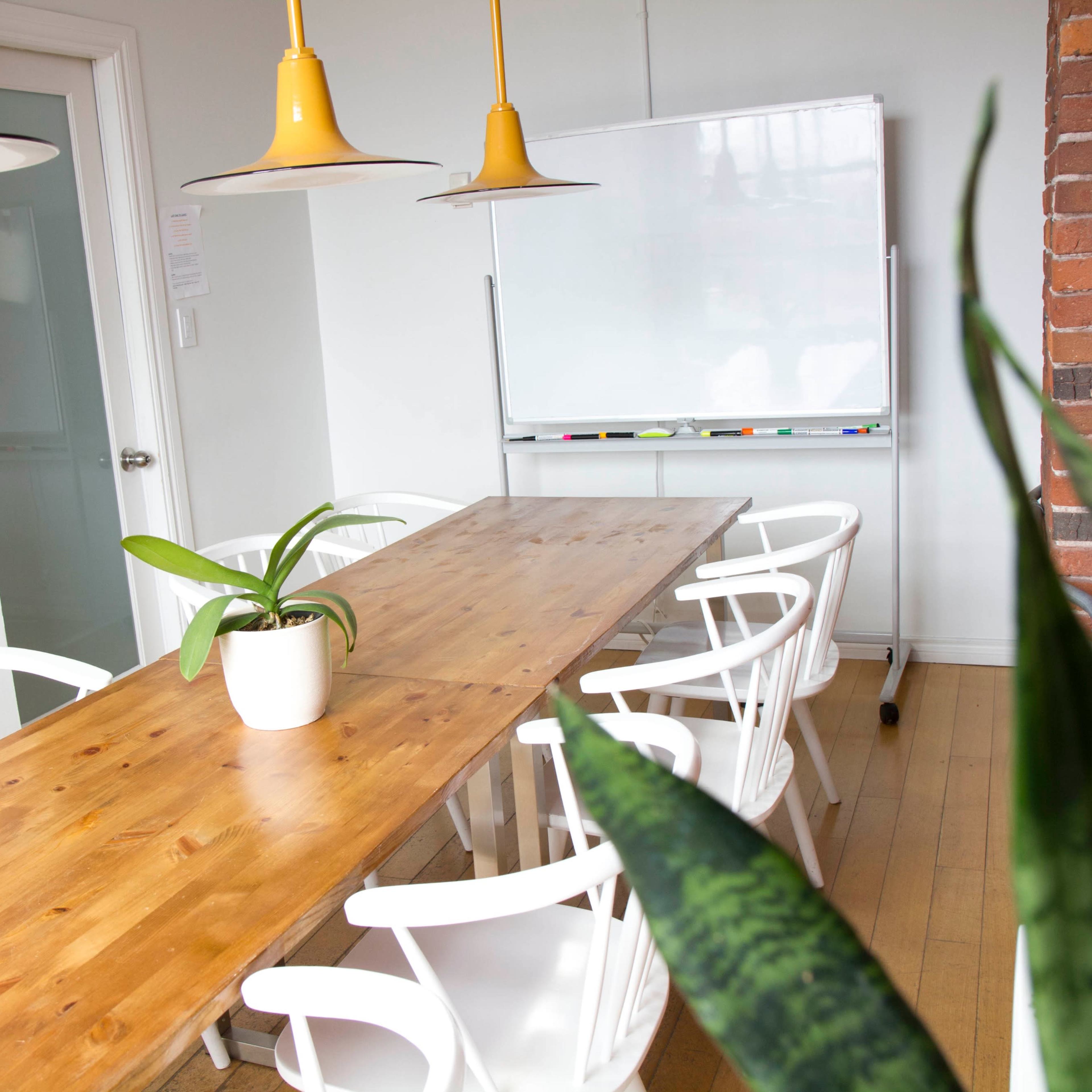 A simple conference room features a long wooden table surrounded by white chairs, with a potted plant and a whiteboard in the background.