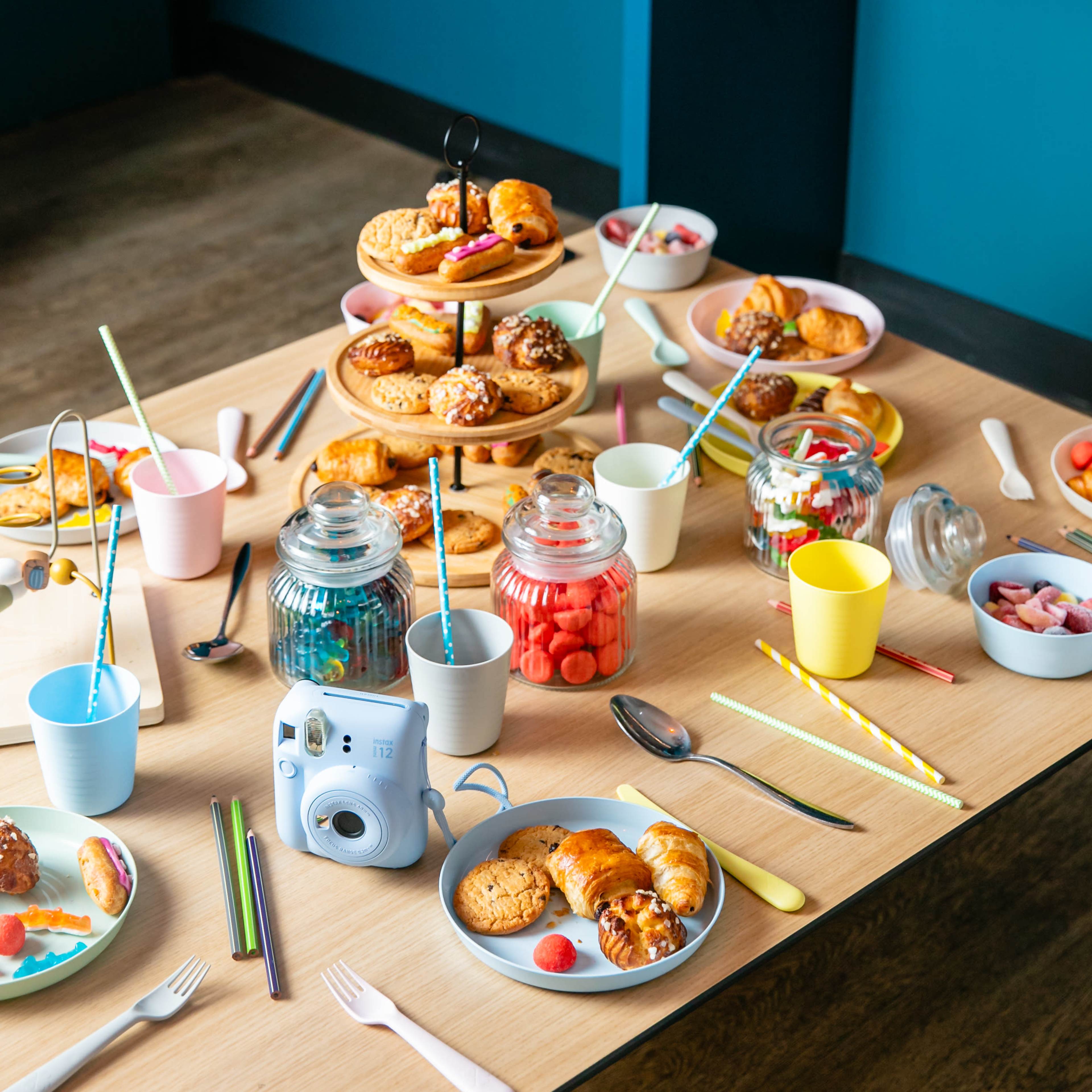 A tabletop is set with a variety of colorful pastries, candies, and beverages, surrounded by plates, cups, and a camera.