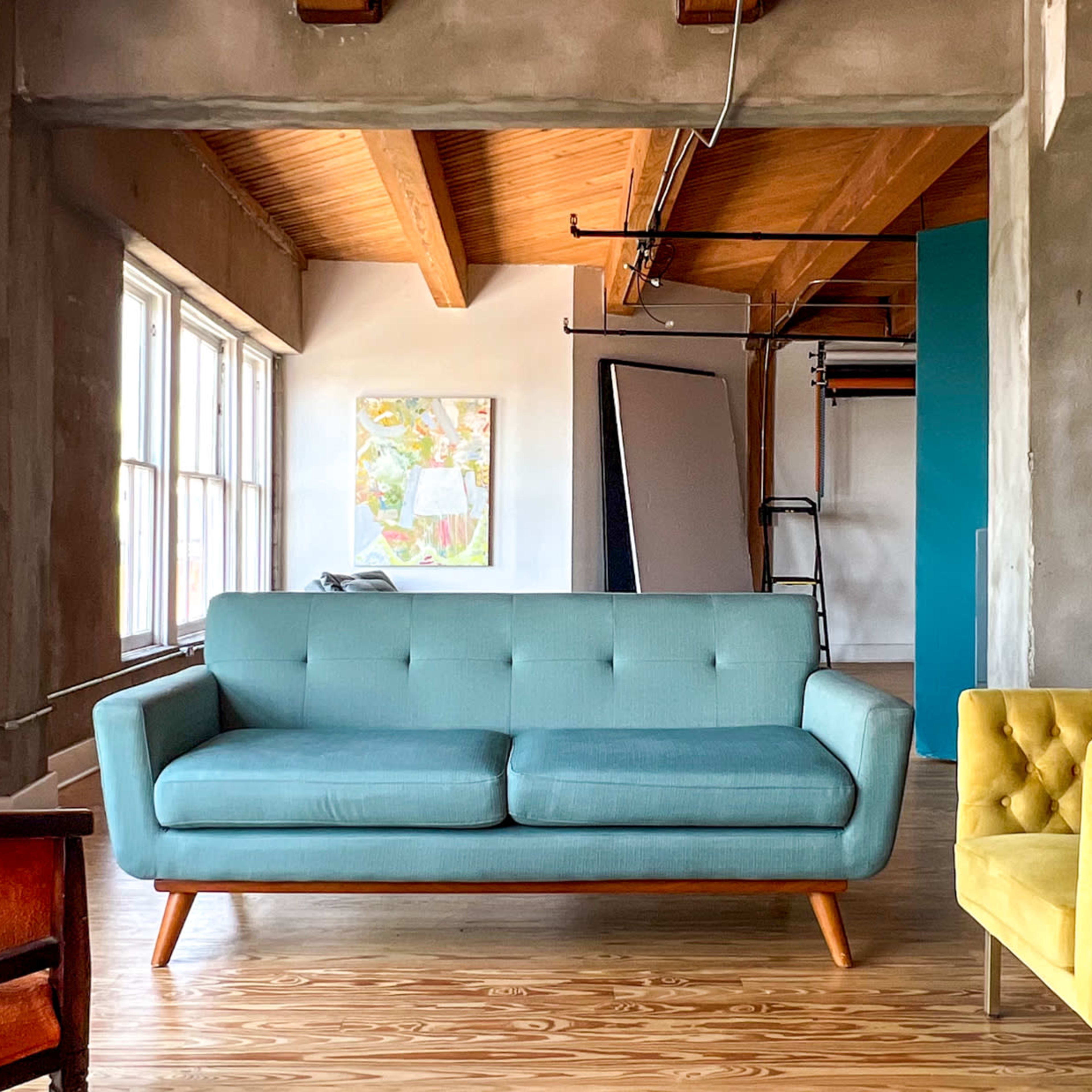 A spacious room featuring a light blue couch, a yellow armchair, and an orange armchair, with large windows and a teal wall in the background.