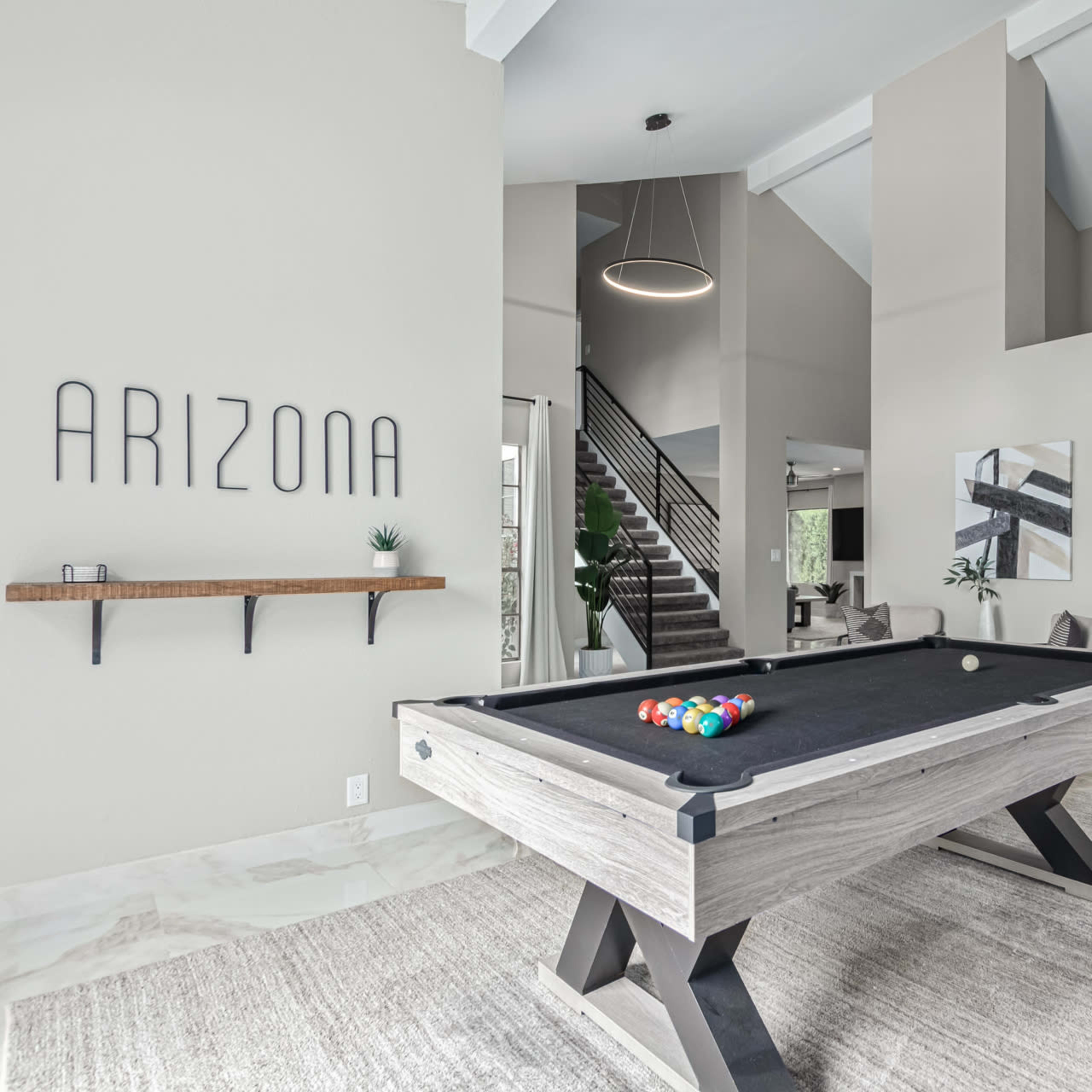 A modern game room features a black pool table, a wall with the word "ARIZONA," and a staircase leading to a second floor.