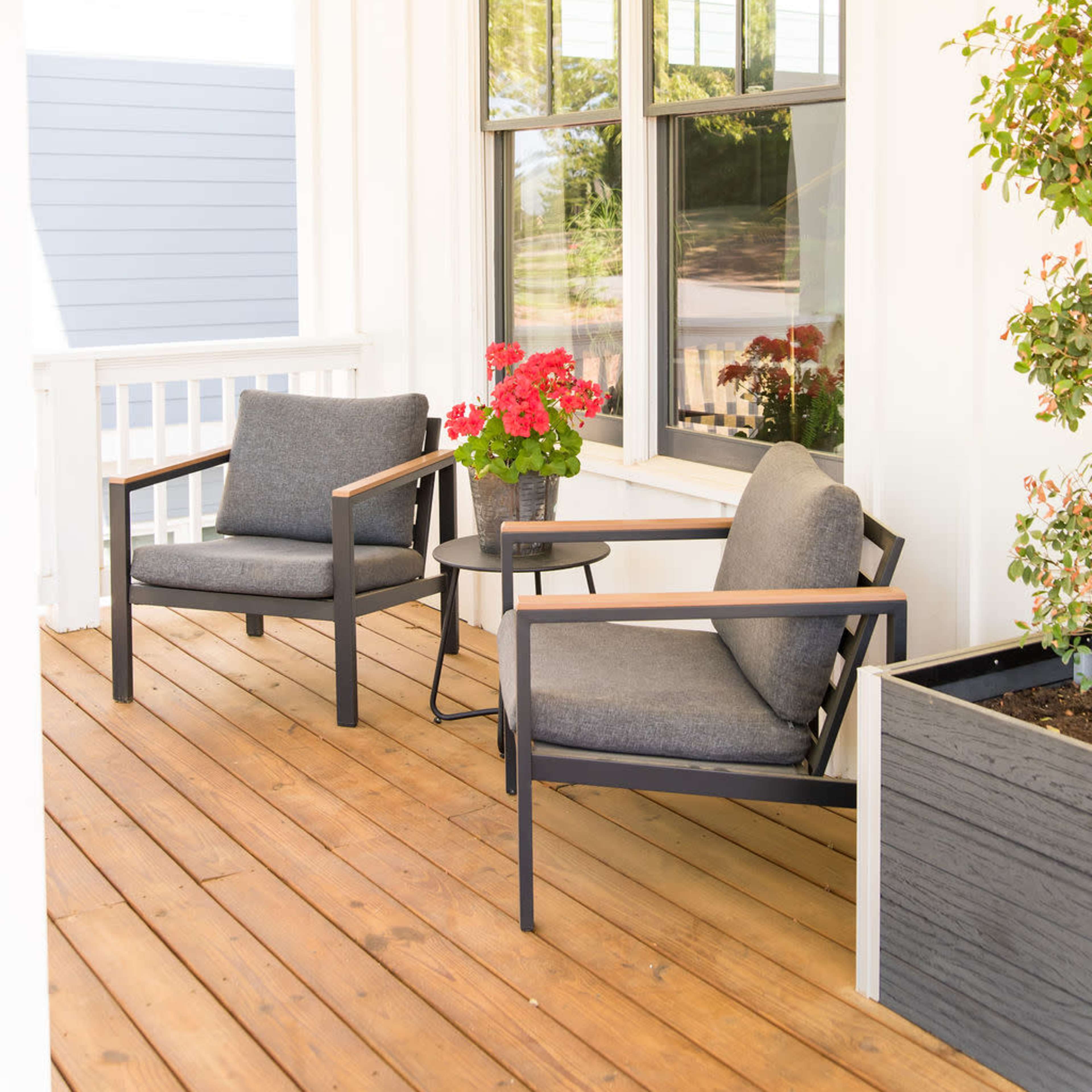 Two gray outdoor chairs beside a small table, positioned on a wooden porch with potted red flowers and large windows in the background.