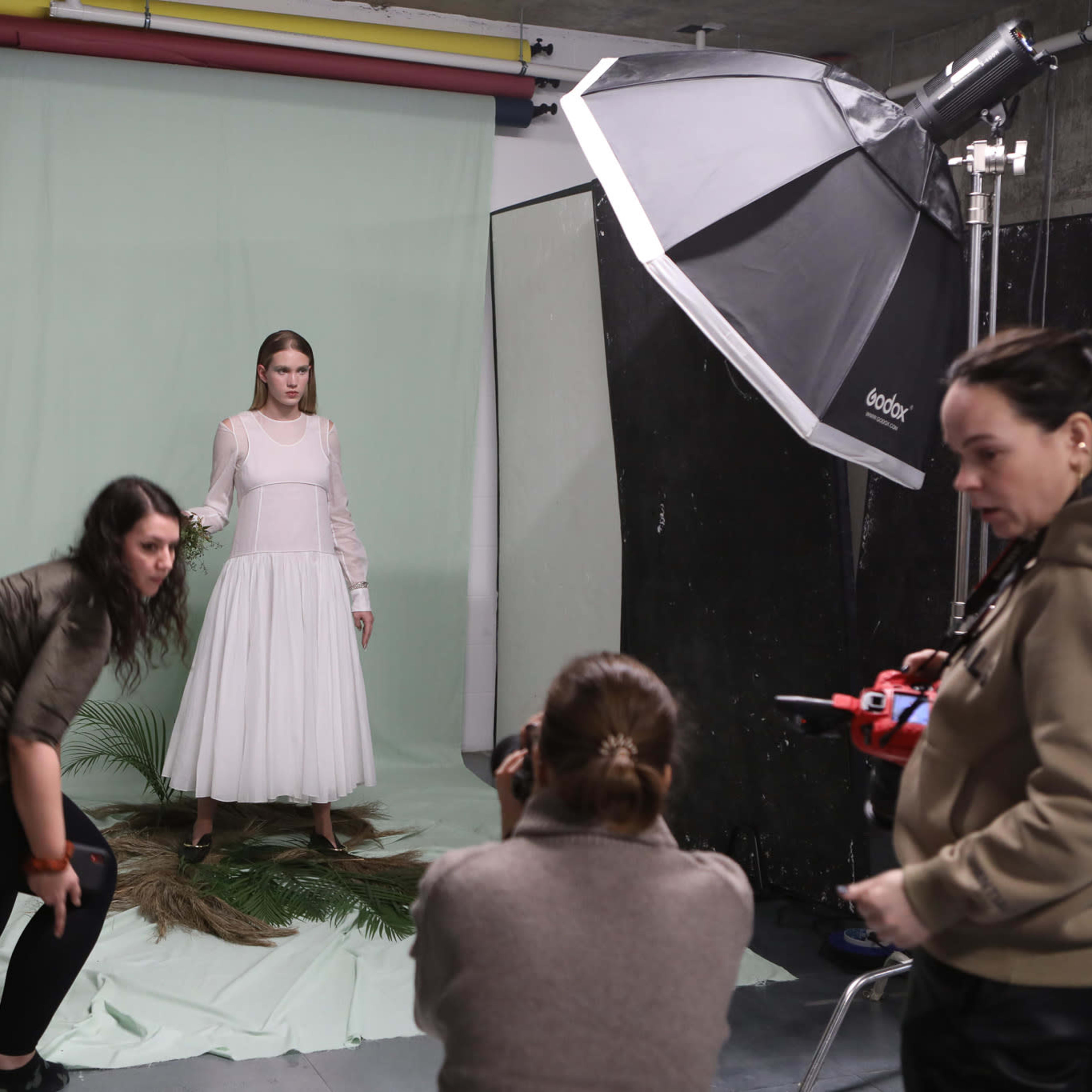 A model stands in a white dress on a green backdrop while two photographers adjust their equipment and take notes.