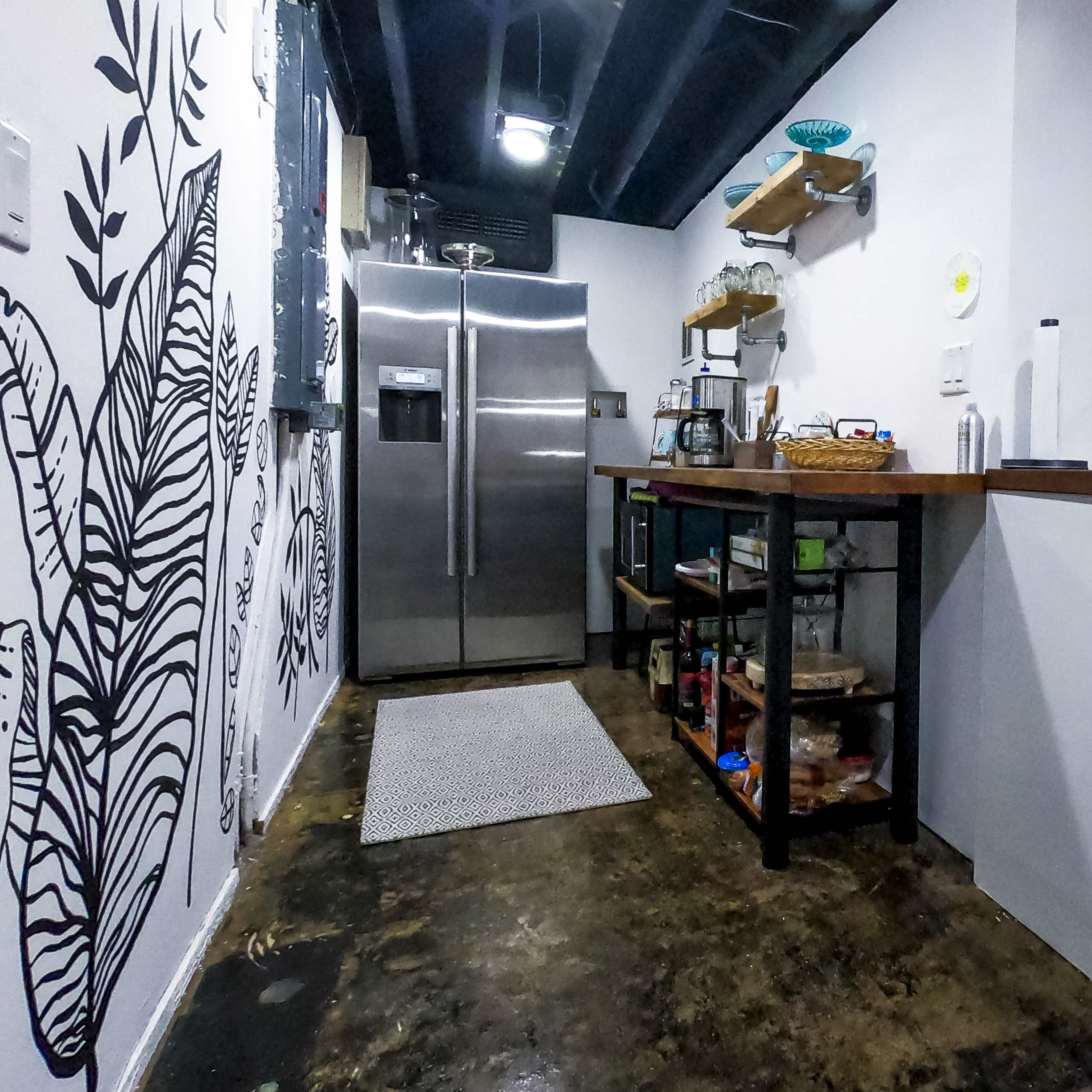A narrow kitchen with a stainless steel refrigerator, a wooden countertop island, and a black-and-white leaf-patterned mural on the wall.