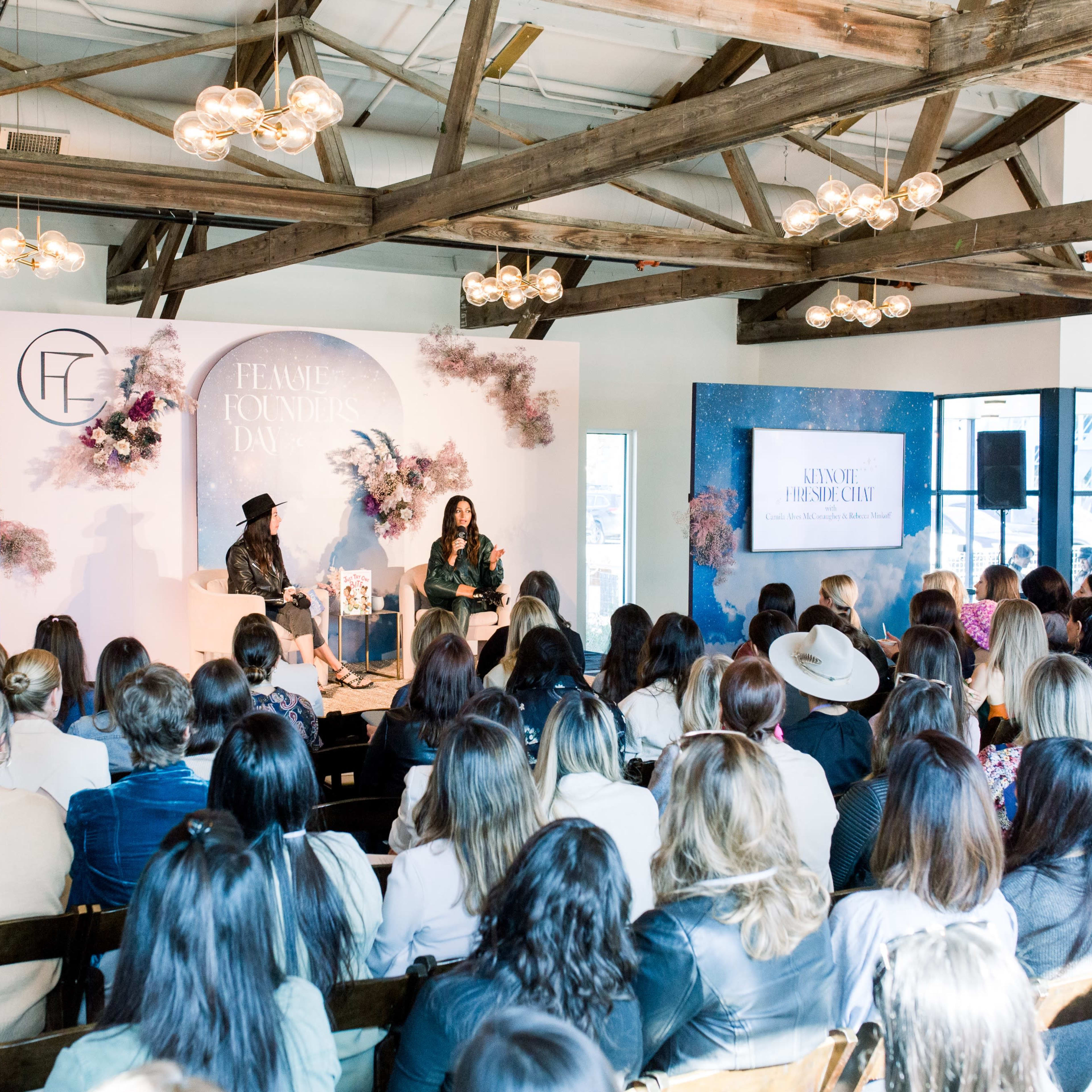 A large audience listens to a speaker at a panel event featuring a decorative stage with floral elements and branding.