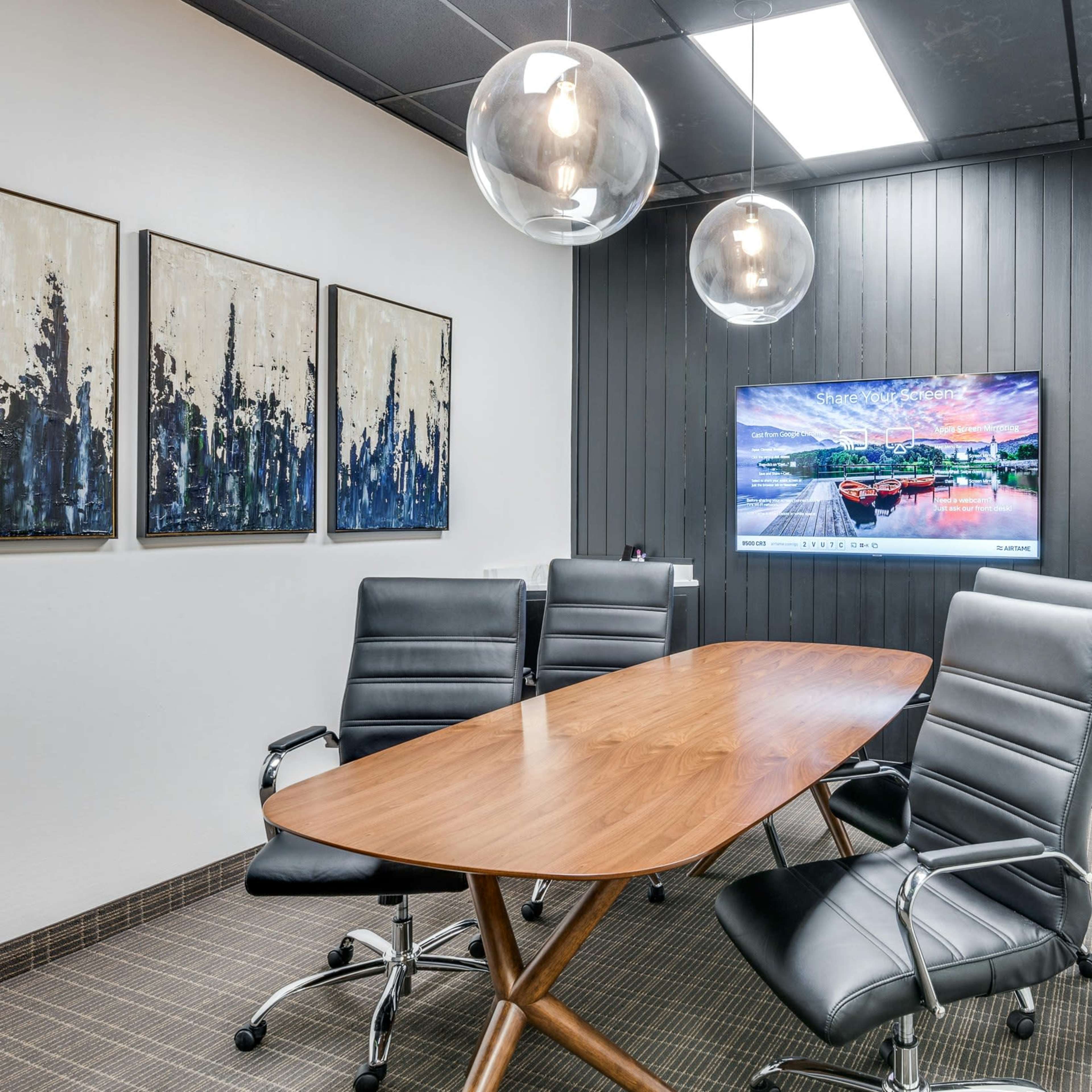 A modern conference room features a wooden table surrounded by black leather chairs, a large wall-mounted TV, and three hanging light fixtures.
