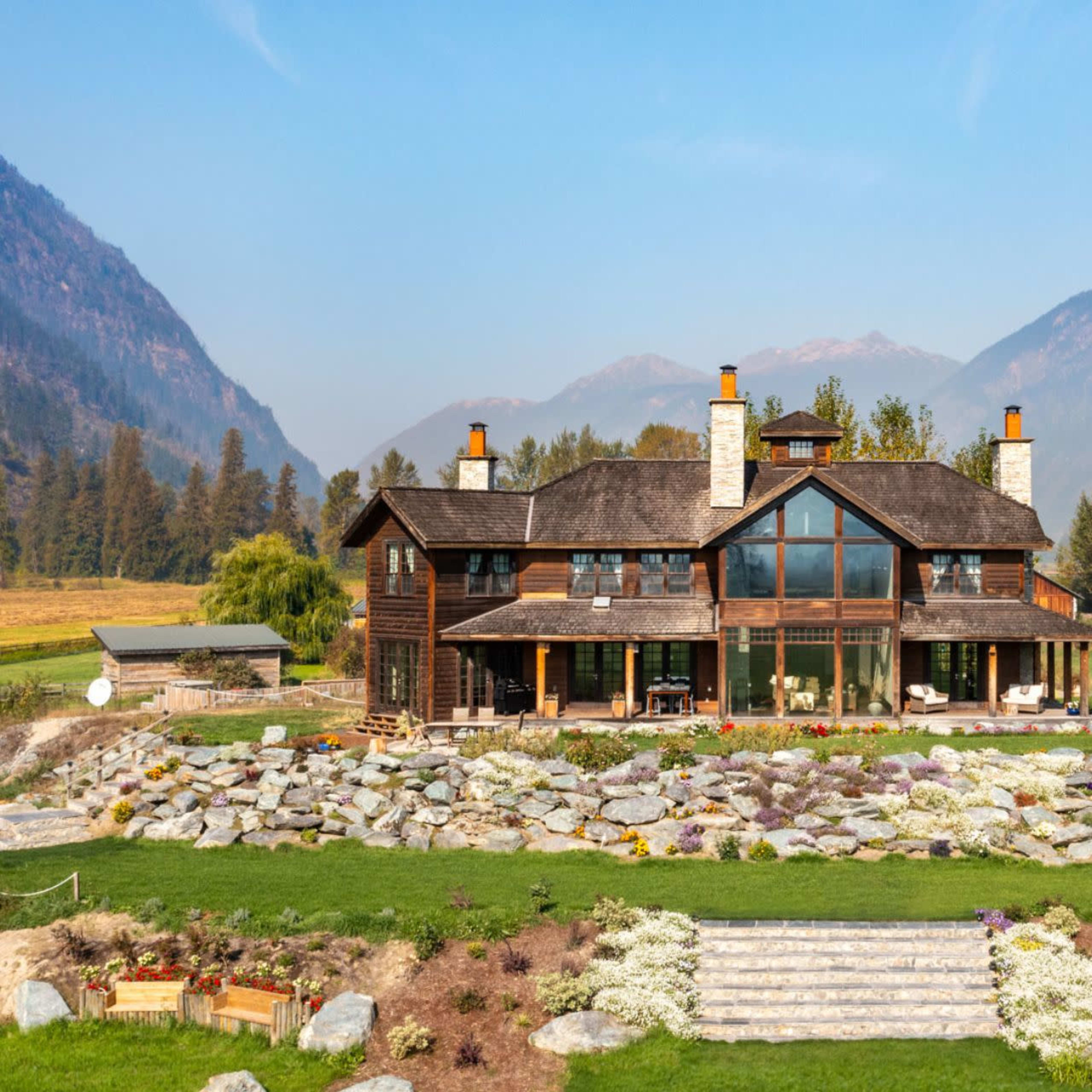 A large wooden house with stone chimneys is set against a backdrop of mountains and a clear blue sky, surrounded by landscaped gardens and rocks.