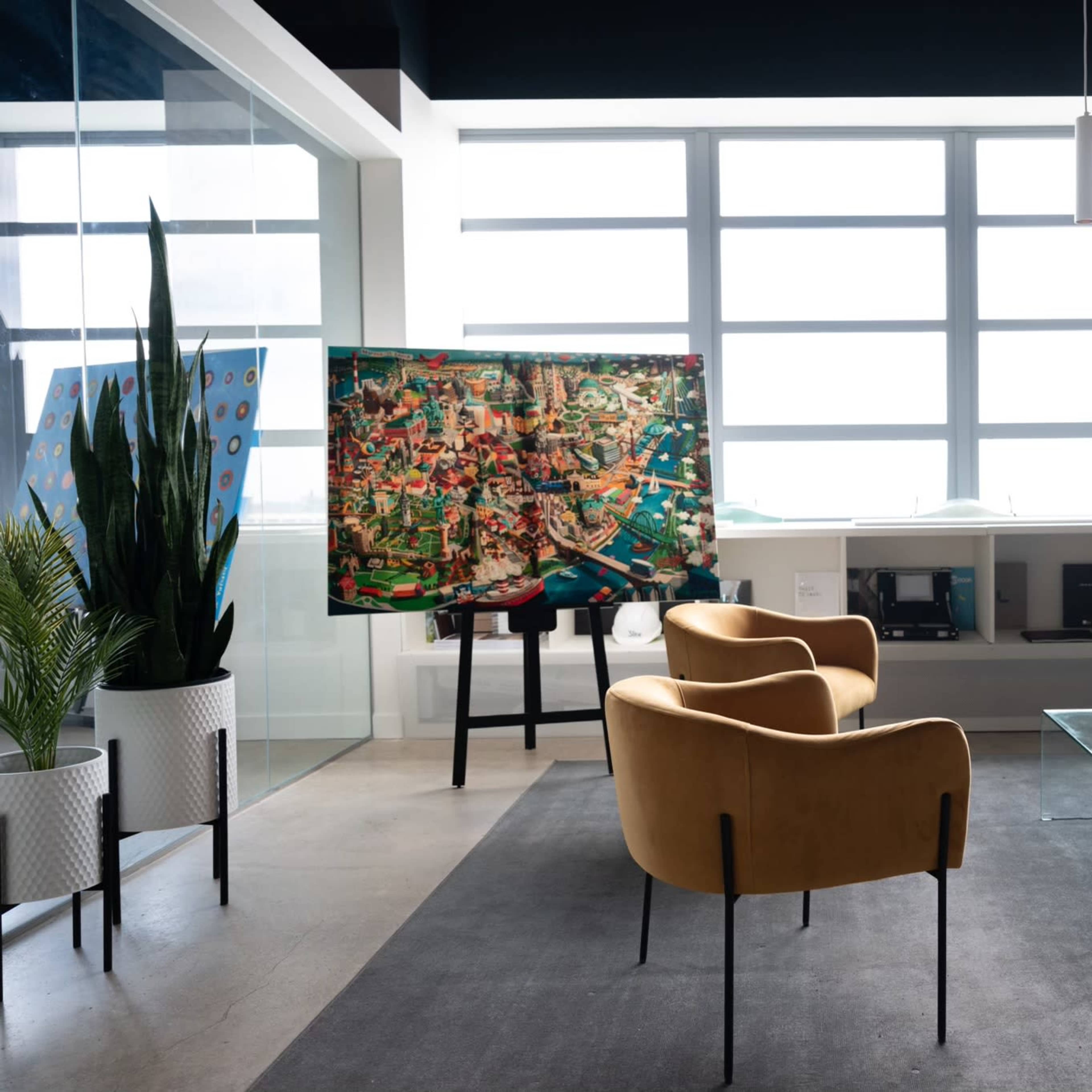 A modern office space featuring two brown chairs, a glass coffee table, and a large colorful artwork displayed on an easel near large windows.