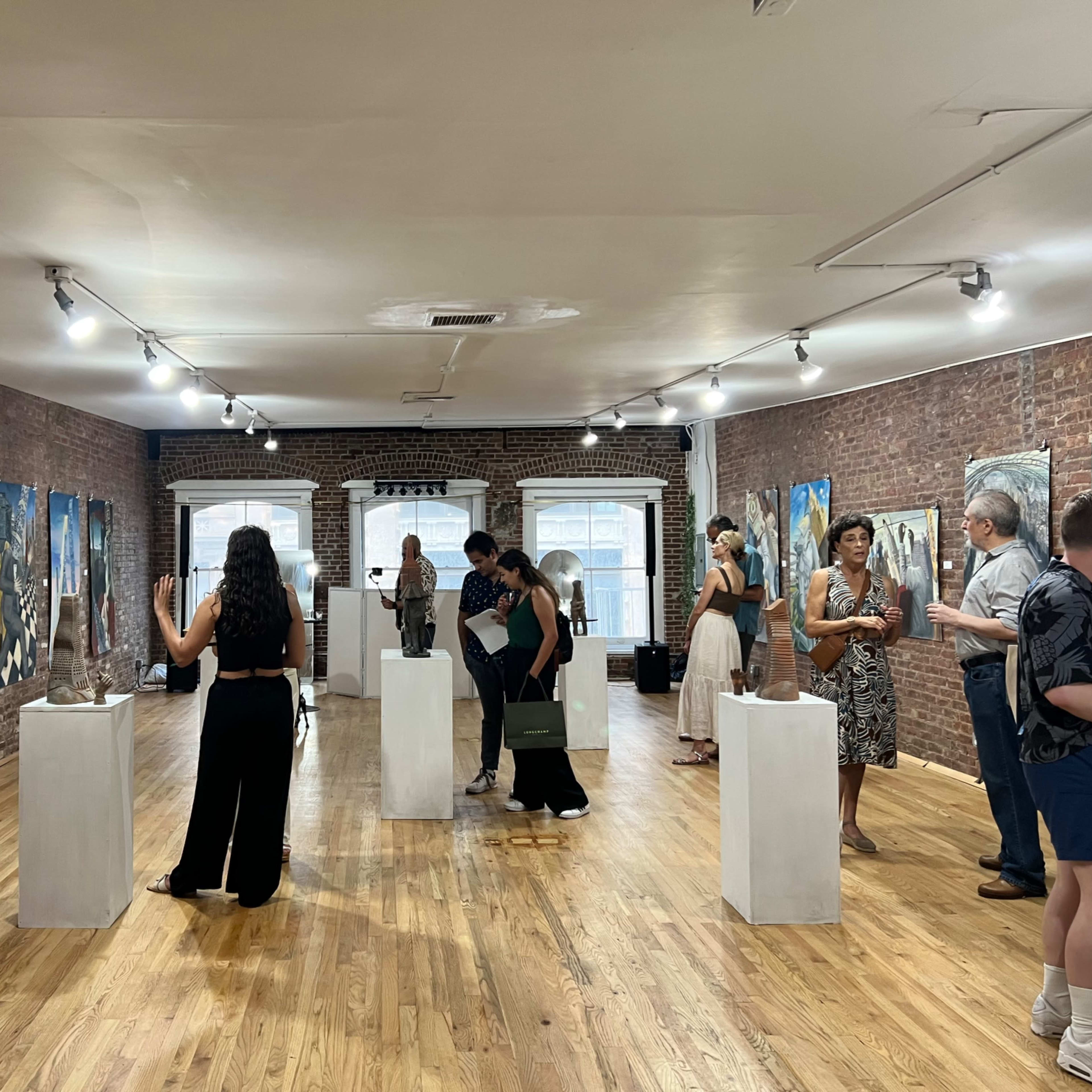A group of people explore an art gallery featuring various paintings and sculptures displayed on pedestals in a room with wooden flooring and brick walls.