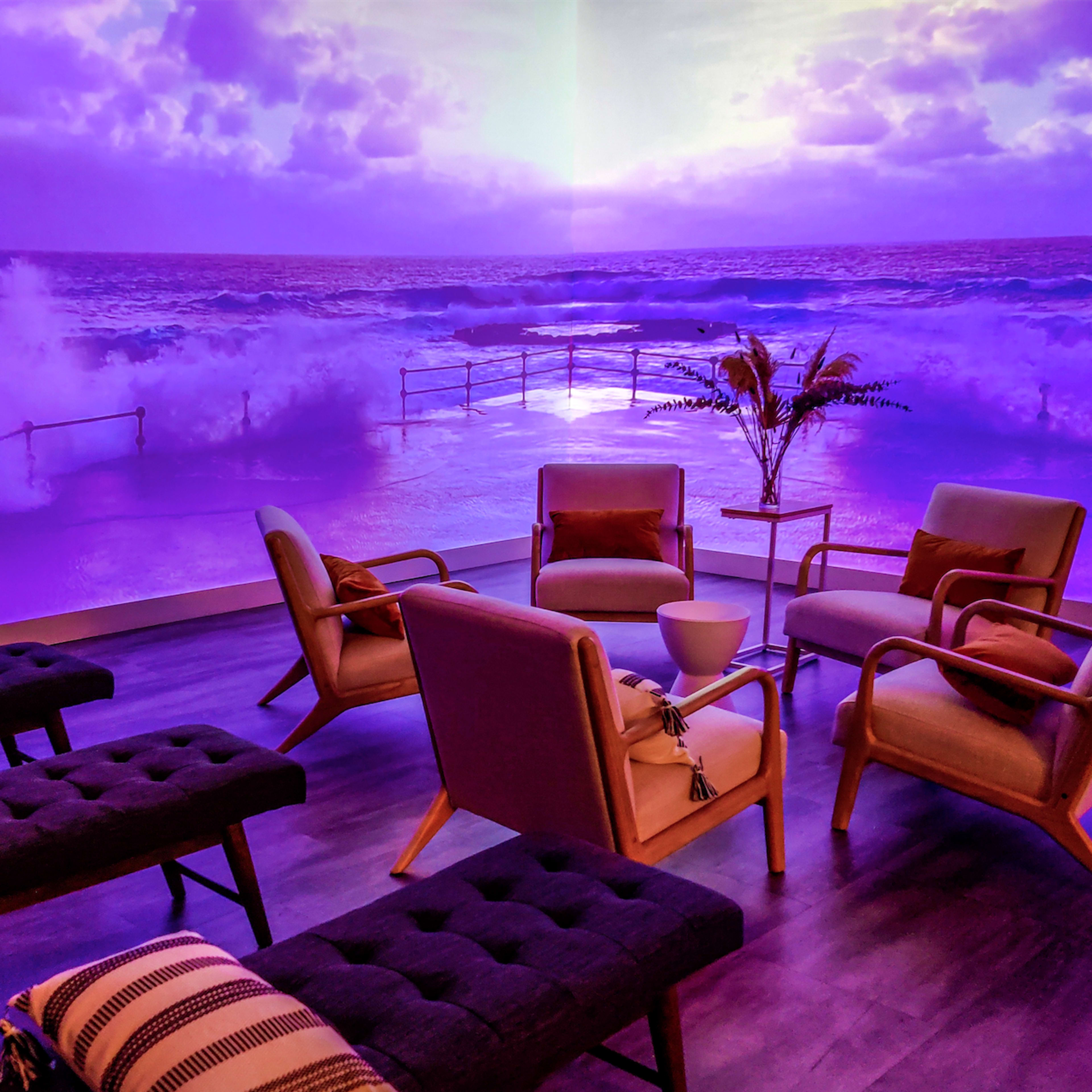 A modern lounge area with several chairs and loungers arranged in front of a large purple-tinted mural of ocean waves.
