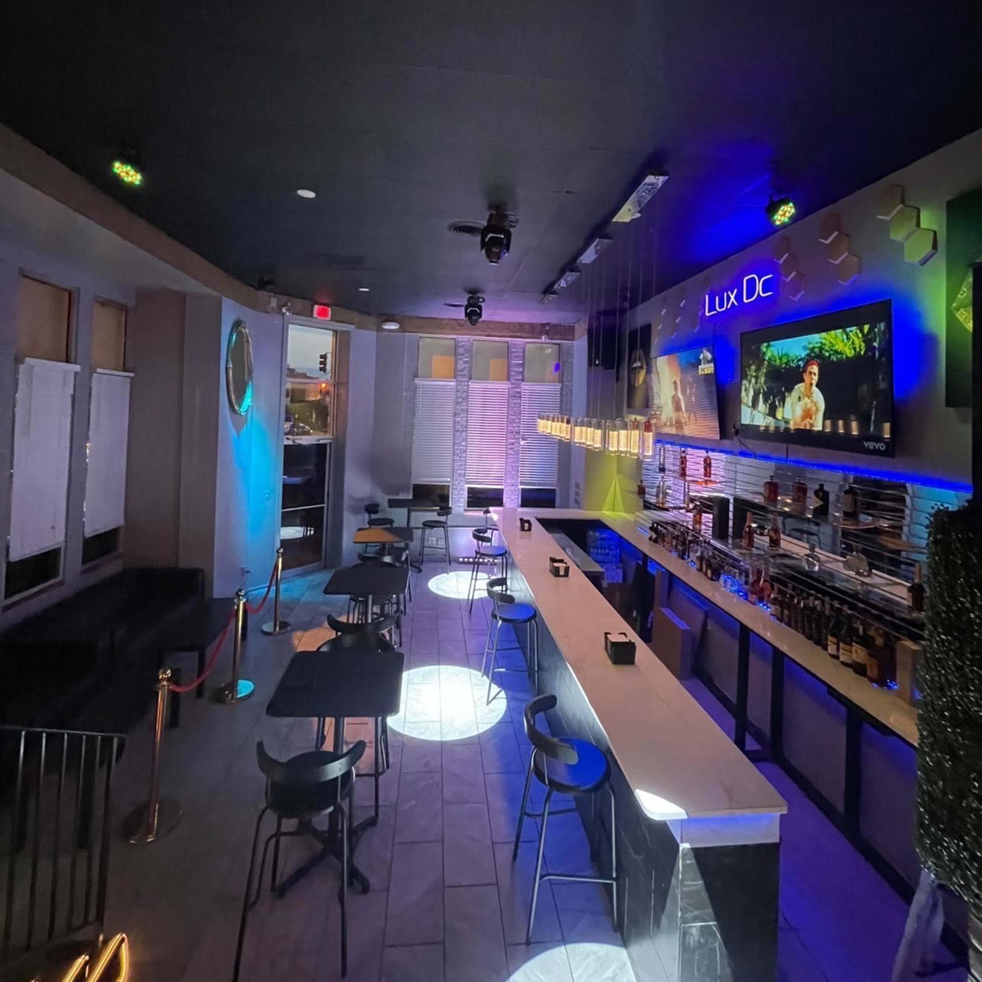 An interior view of a bar with a long counter, high chairs, and ambient lighting, featuring a television mounted on the wall.