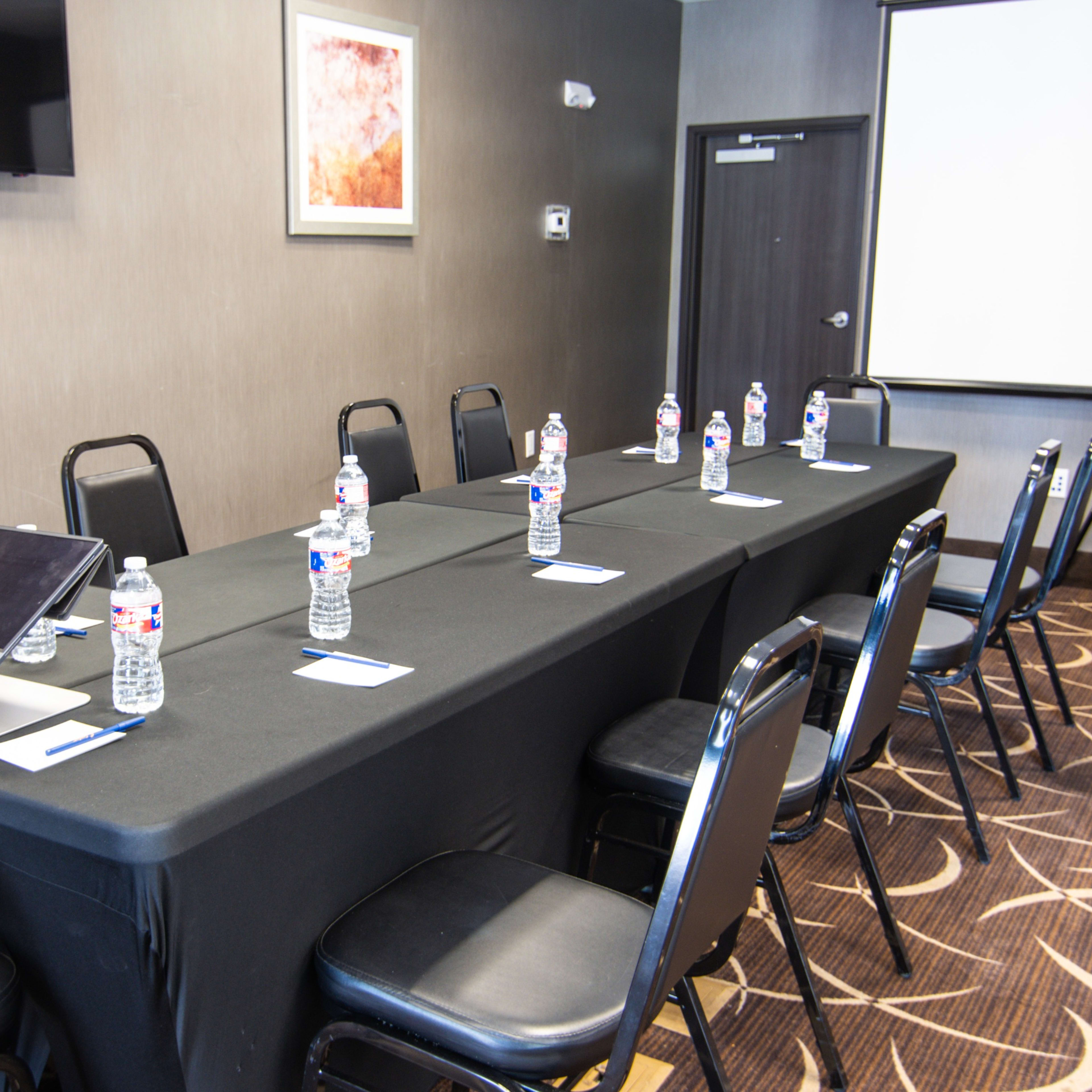 A conference room setup features a long table with chairs, each equipped with a bottle of water and a name tag, alongside a laptop and a screen for presentations.