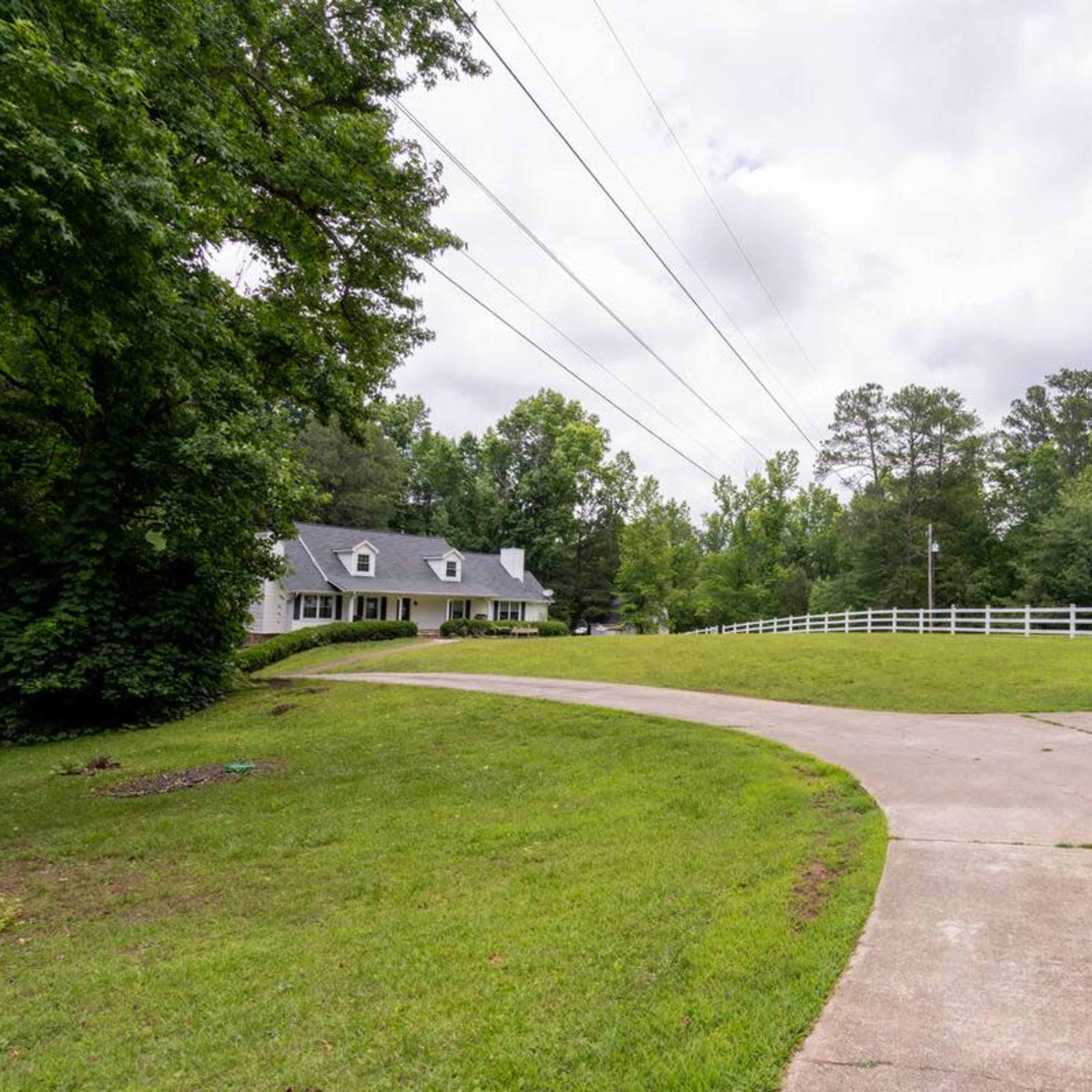 A gravel driveway curves past a house surrounded by greenery and a white fence.