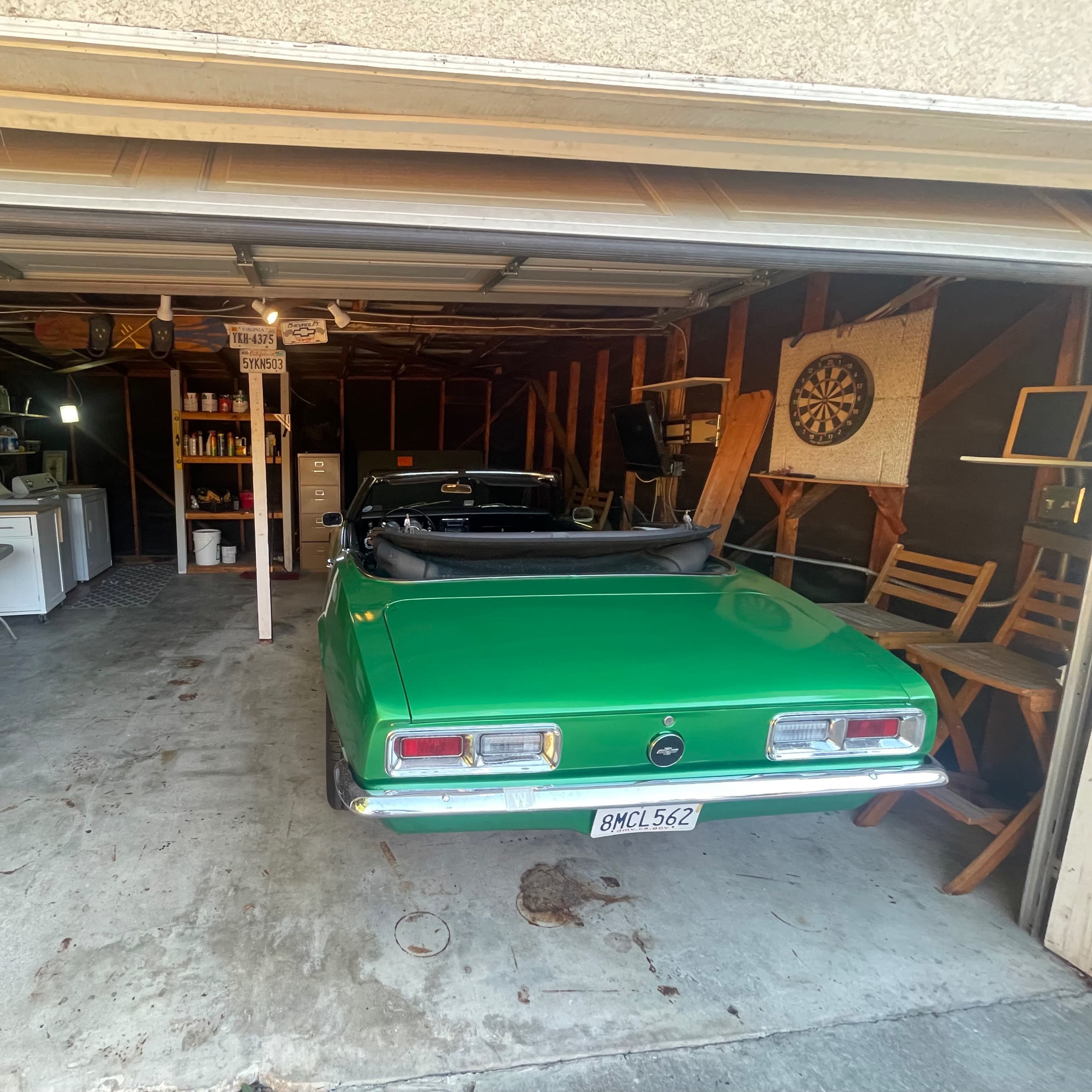 A green convertible car is parked in a garage that also contains a small table, chairs, and various storage items.