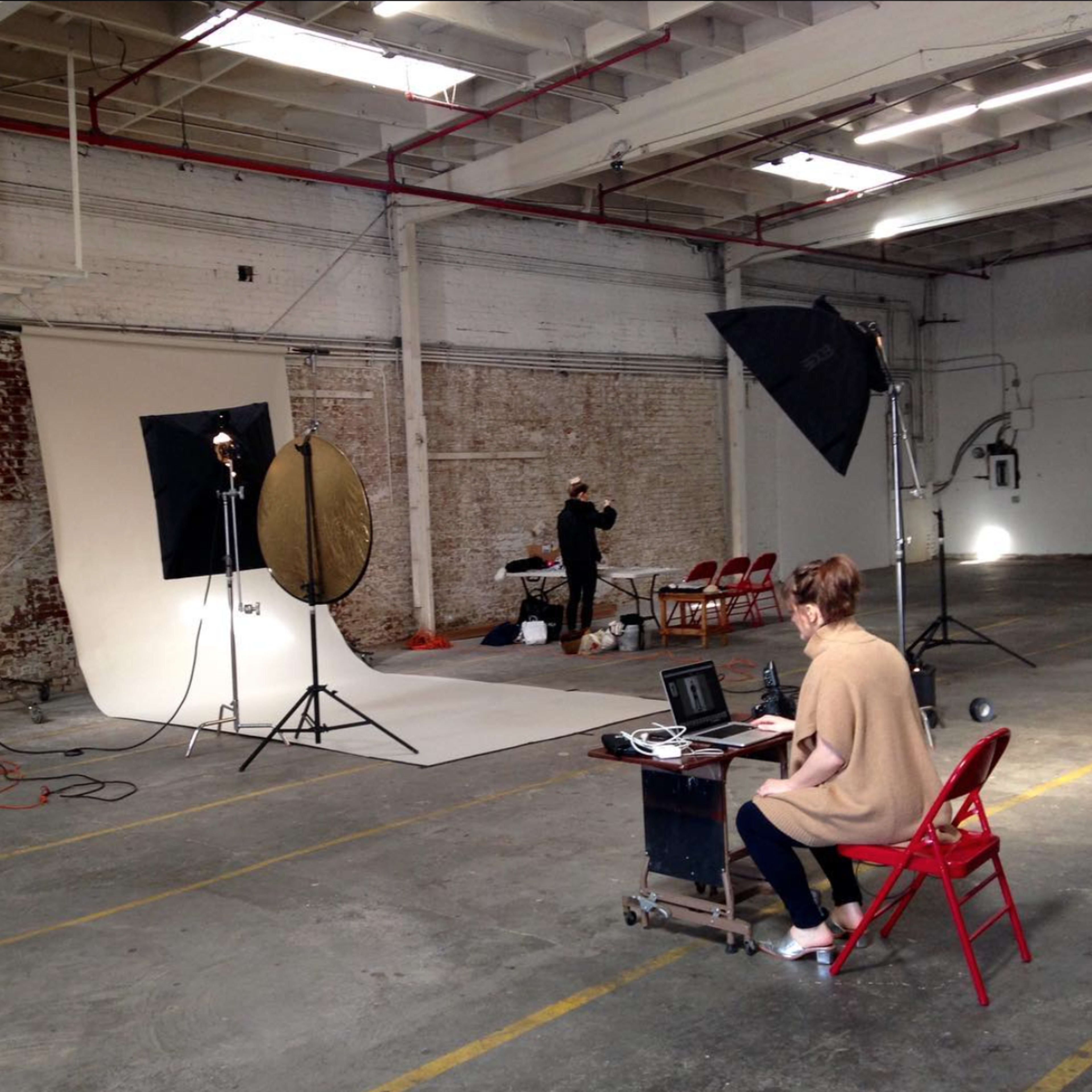 A photographer sets up equipment in an empty studio with a model seated in front of a backdrop, while a person operates a laptop nearby.