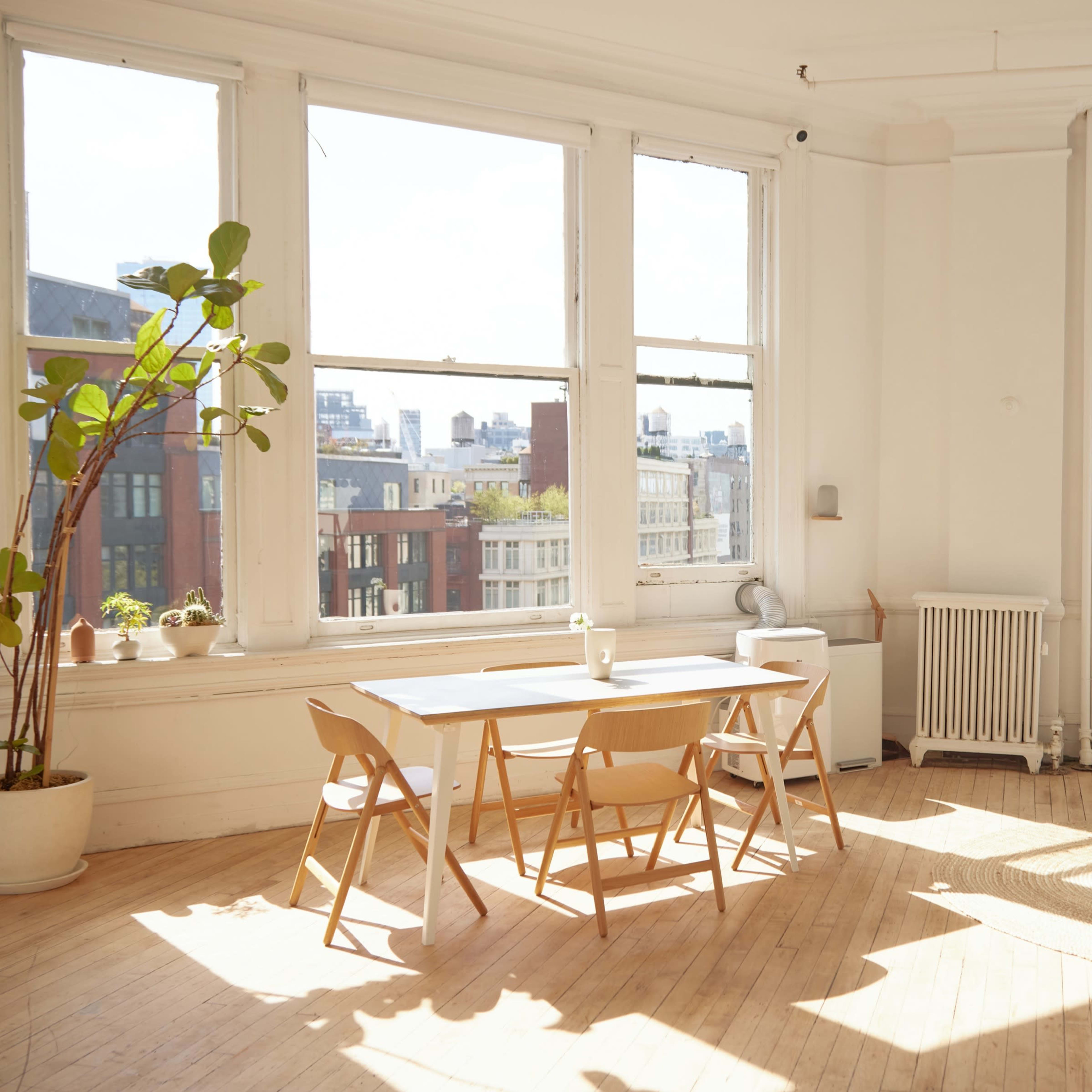 A bright, airy room with large windows, a wooden dining table with chairs, and a light-colored sofa, all set on a wooden floor.
